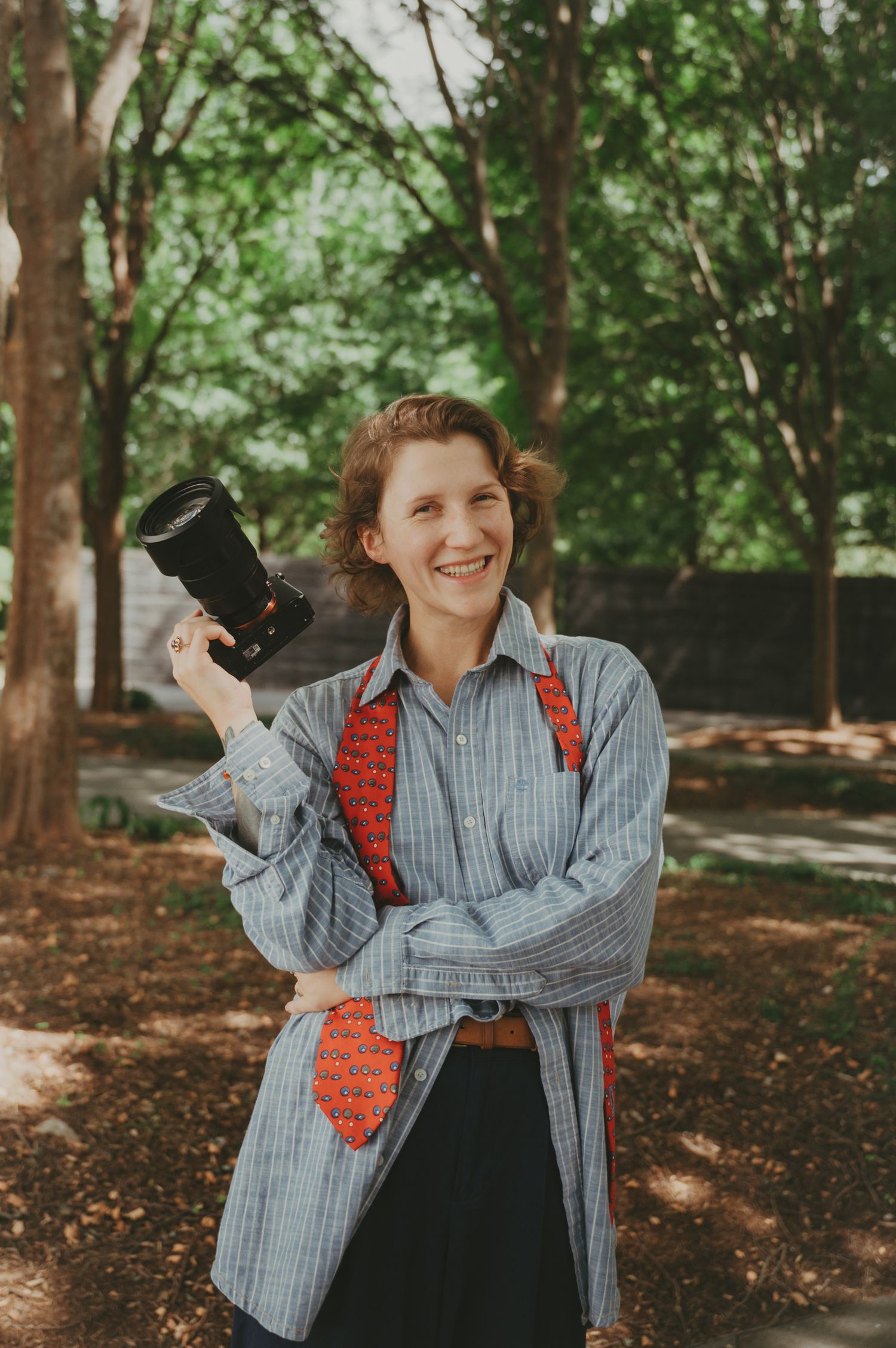 Smiling woman holding a camera outdoors, wearing a striped shirt and red tie, surrounded by green trees.