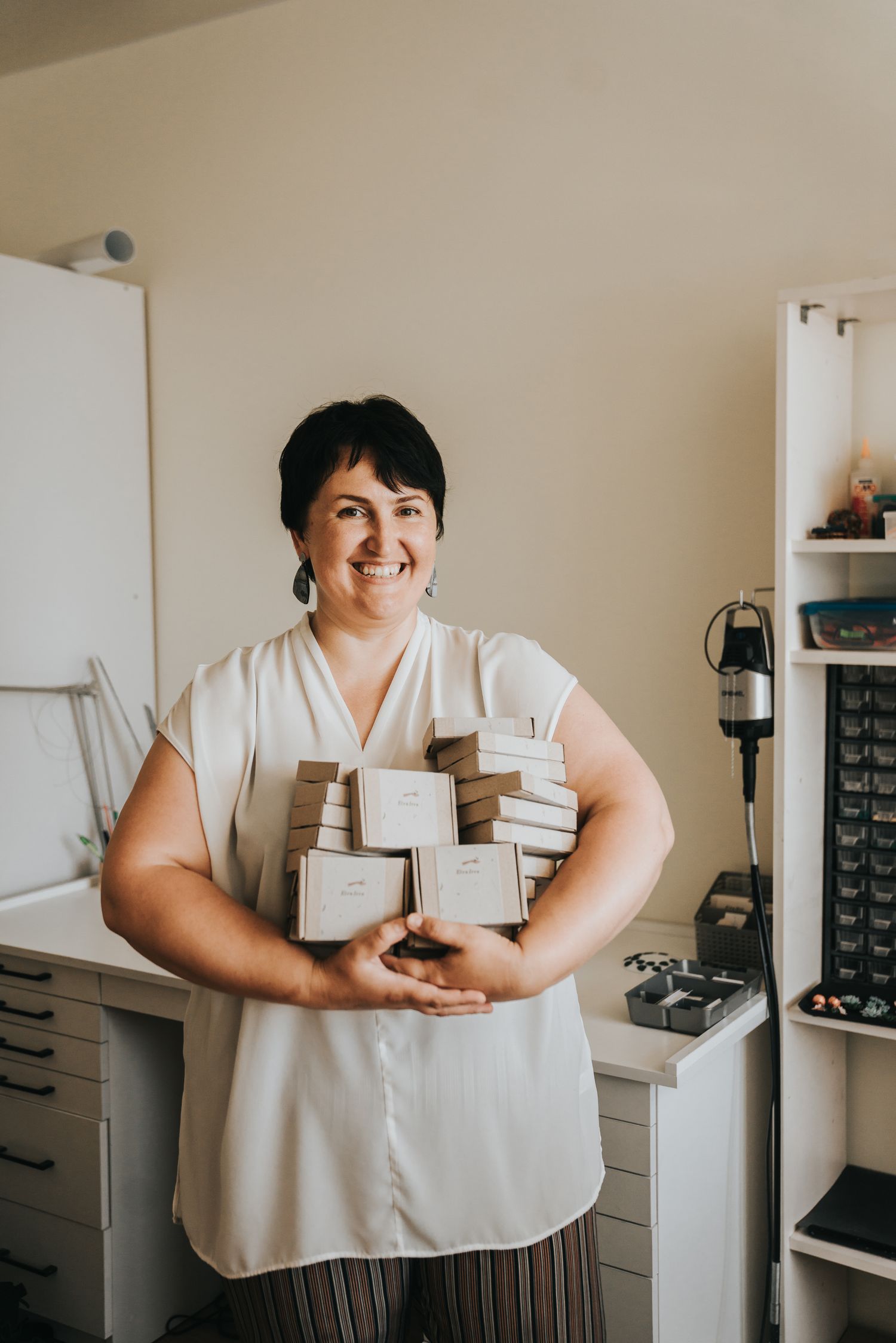 Smiling woman holding a stack of kraft gift boxes in a bright home office workspace.
