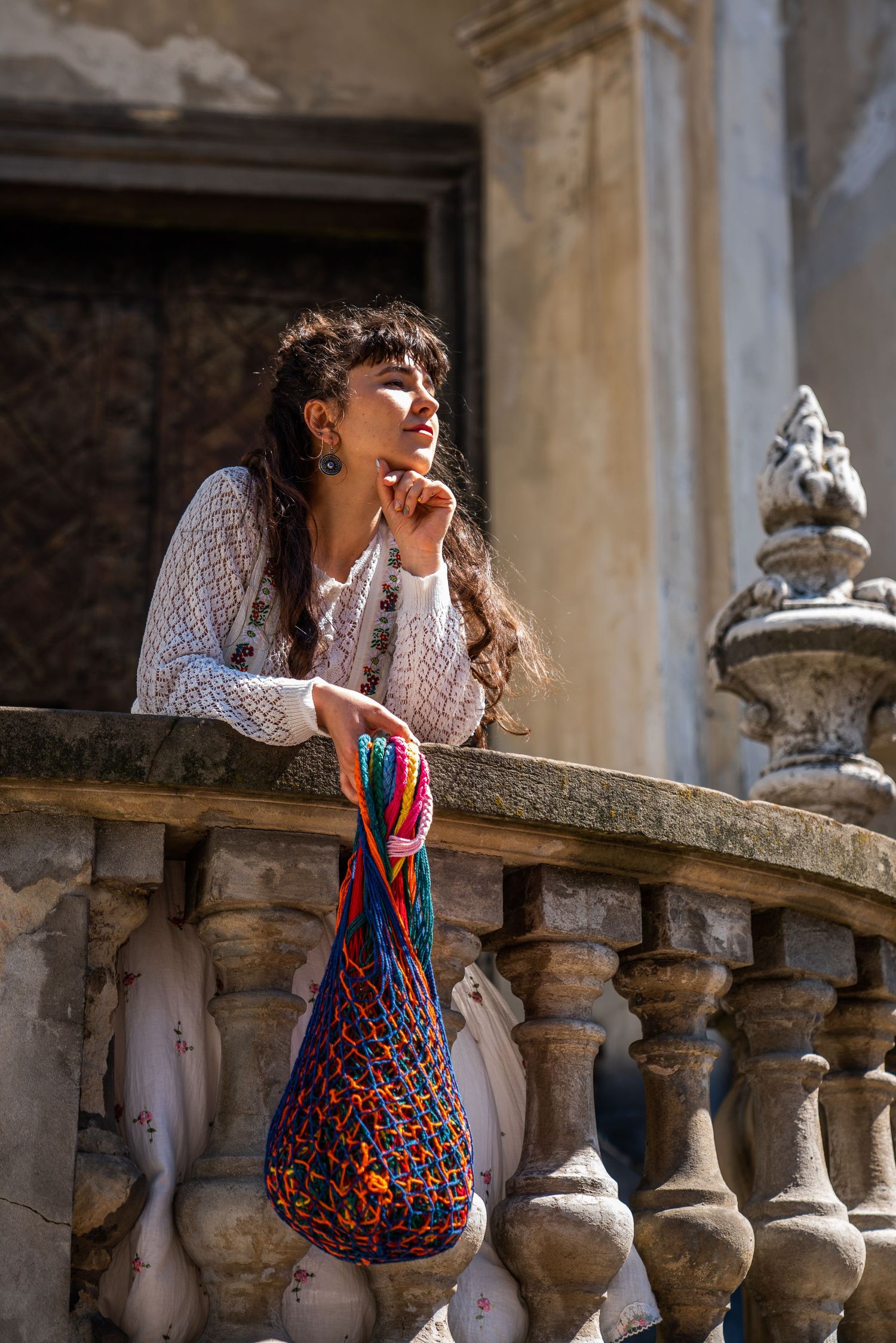 Young woman leaning on ornate stone balcony railing, holding colorful mesh bag, wearing white lace top in sunlight.