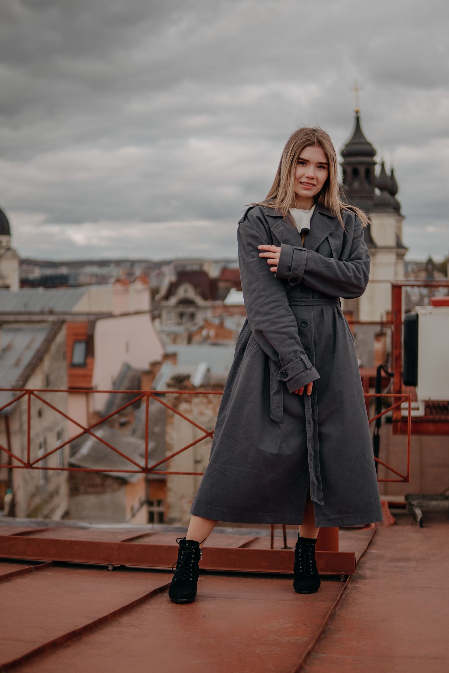 Young blonde woman in gray trench coat and black boots poses on rooftop with European city skyline backdrop.