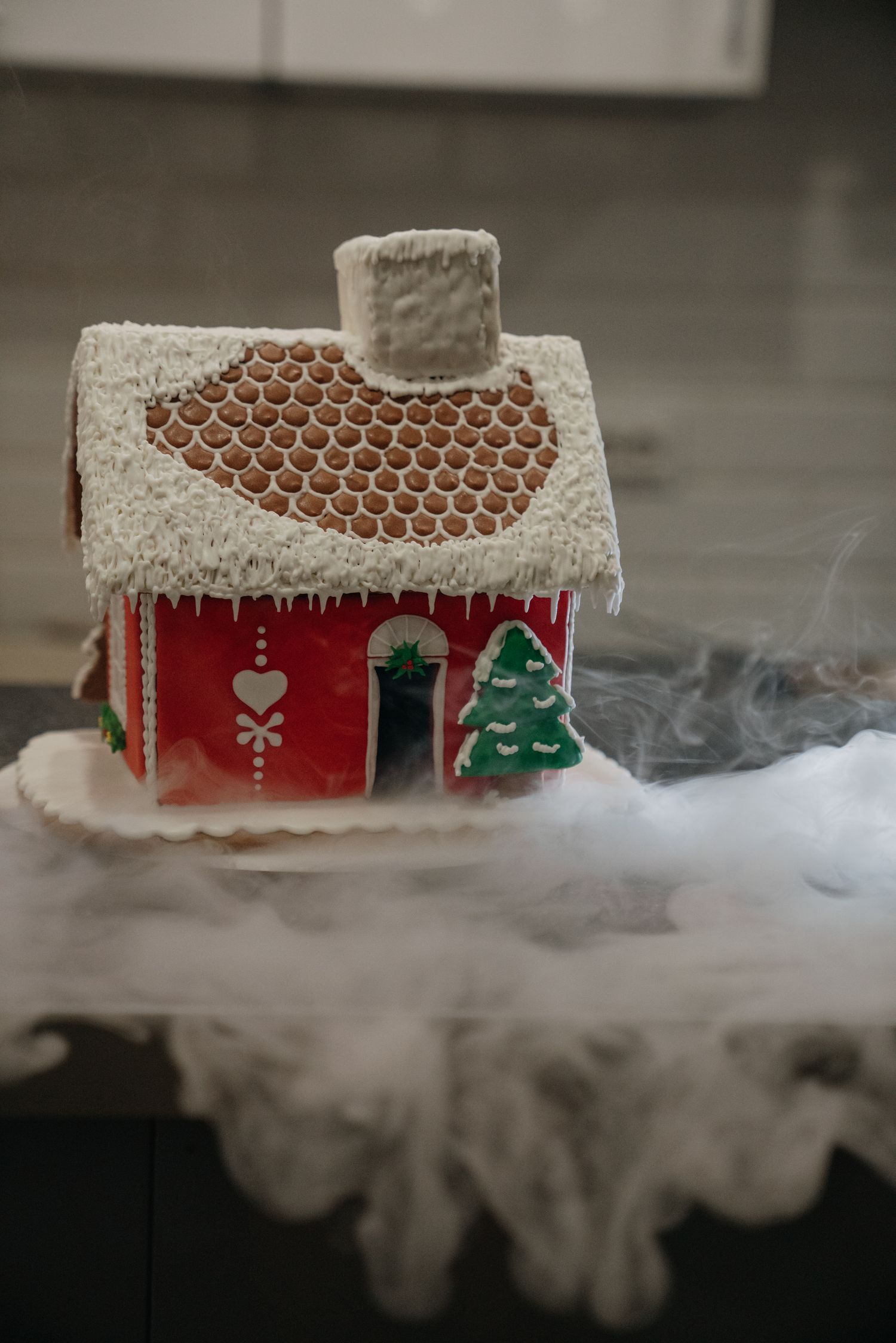 A festive red gingerbread house with white icing, Christmas tree decor, and dry ice fog effect on a dark surface.