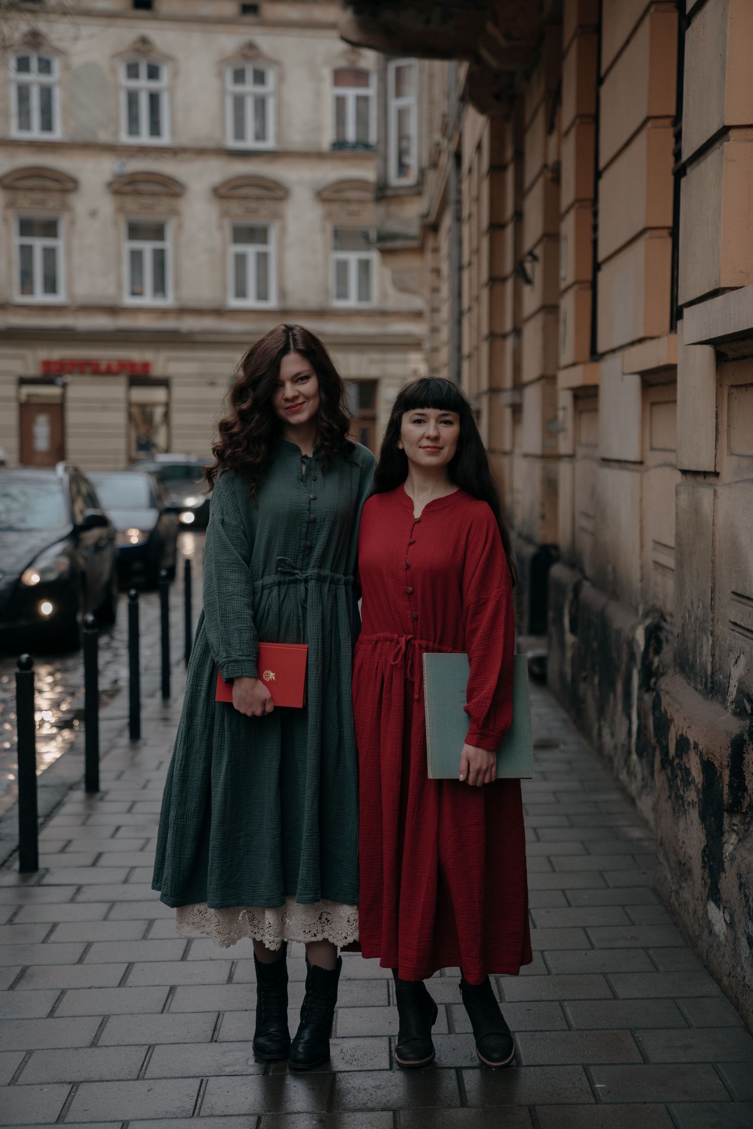 Two women in elegant green and red midi dresses holding books pose on a cobblestone European city street.