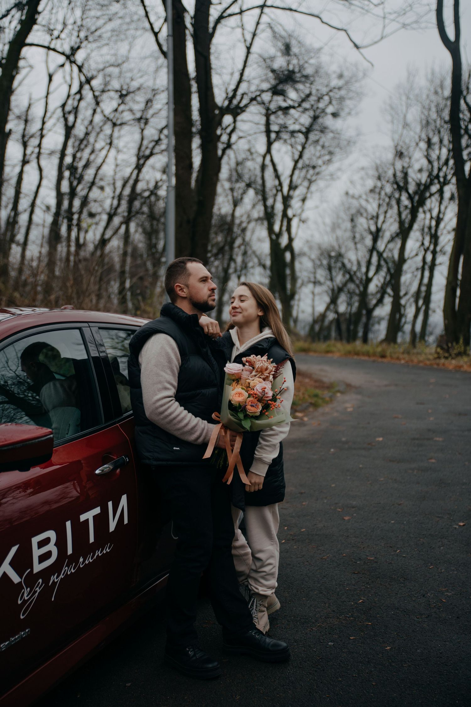 Couple embracing beside a red car on a tree-lined road, woman holding small bouquet.