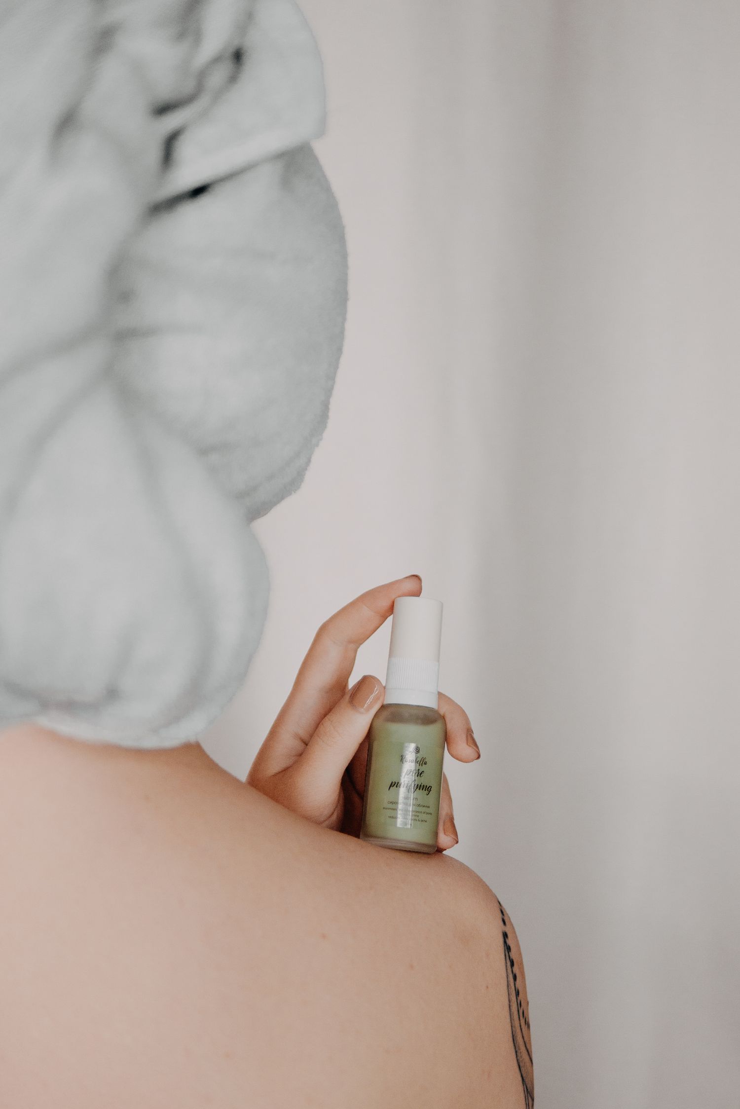 Woman with towel wrapped hair holding a green skincare serum bottle, showing a shoulder tattoo against white background.