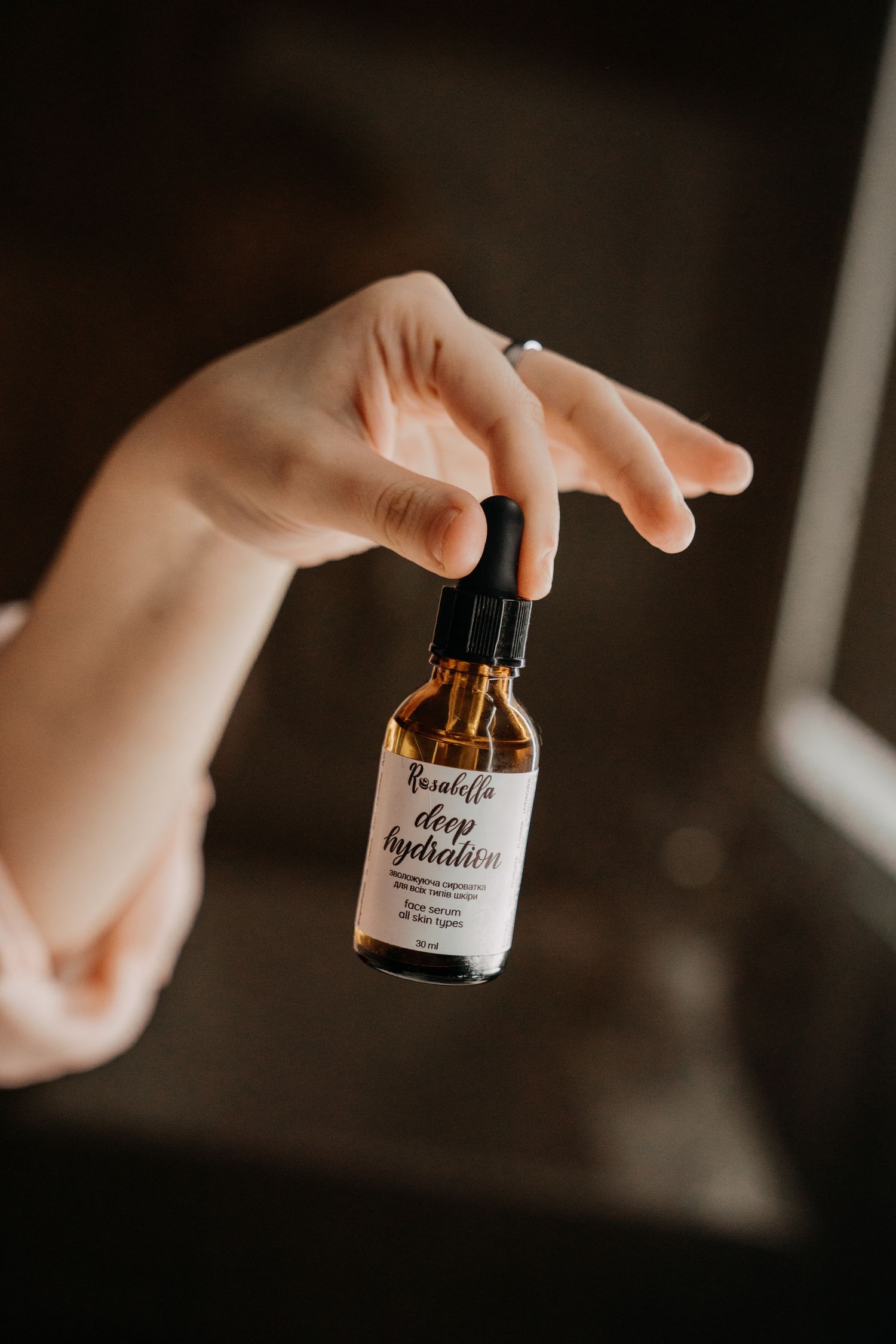 Hand holding a Rosabella Deep Hydration facial serum in an amber dropper bottle.