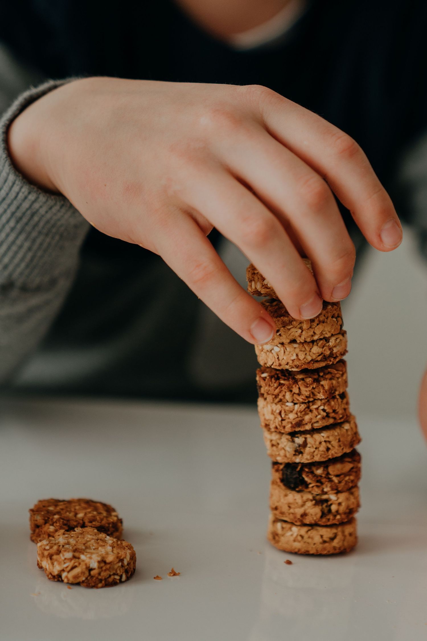 Child's hand stacking oat granola cookies into a tall tower on a white reflective surface.
