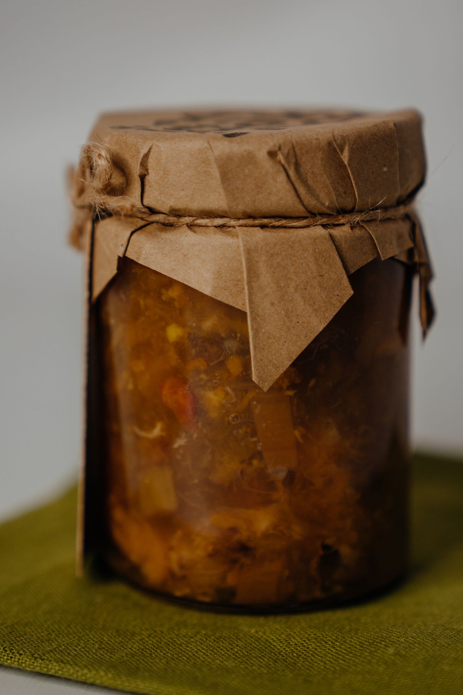 A rustic glass jar of homemade vegetable preserve sealed with brown kraft paper and twine, placed on a green cloth.