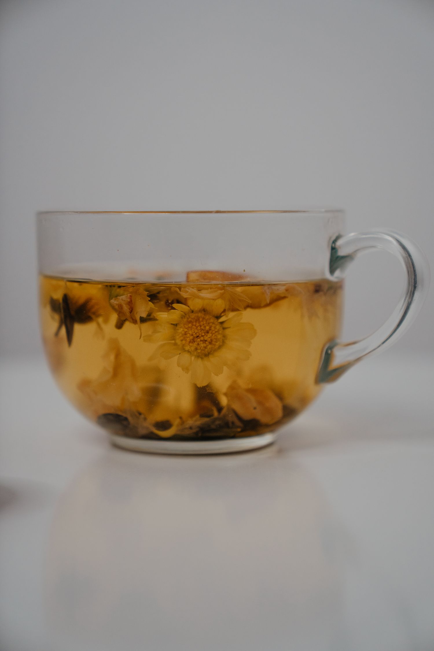 Clear glass cup filled with golden chamomile flower herbal tea on a white surface.