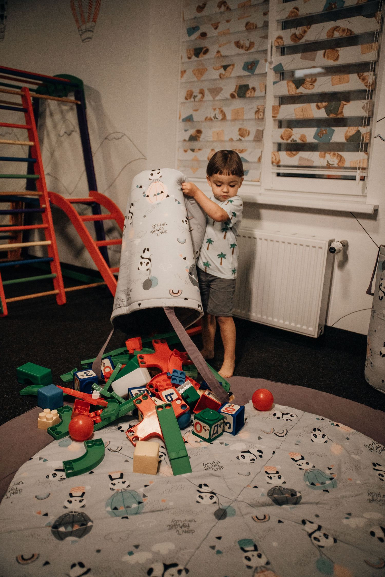 Young boy emptying a toy bag onto a playmat surrounded by colorful wooden blocks.