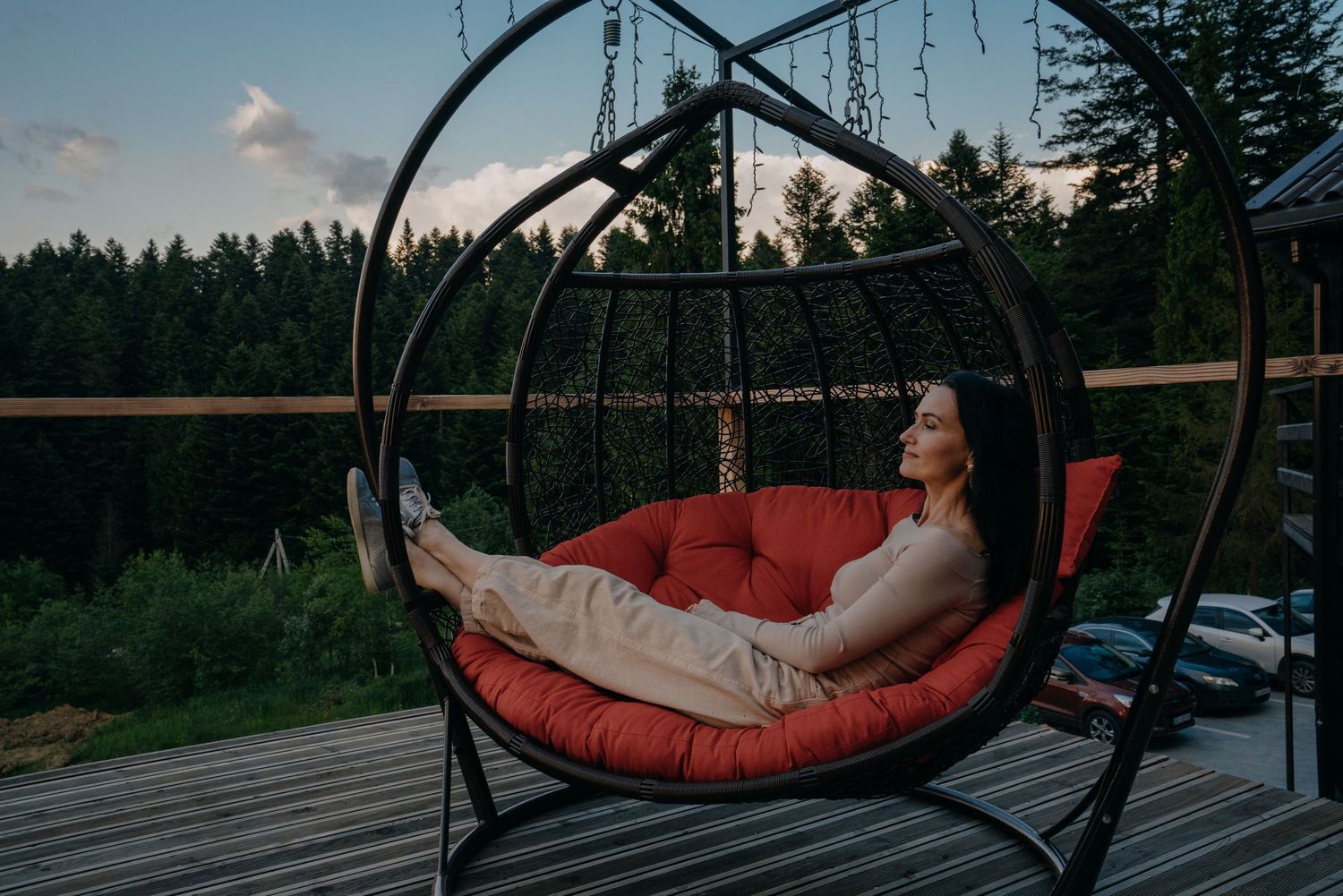Woman relaxing in a hanging egg chair with red cushion on a wooden deck surrounded by lush forest at dusk.