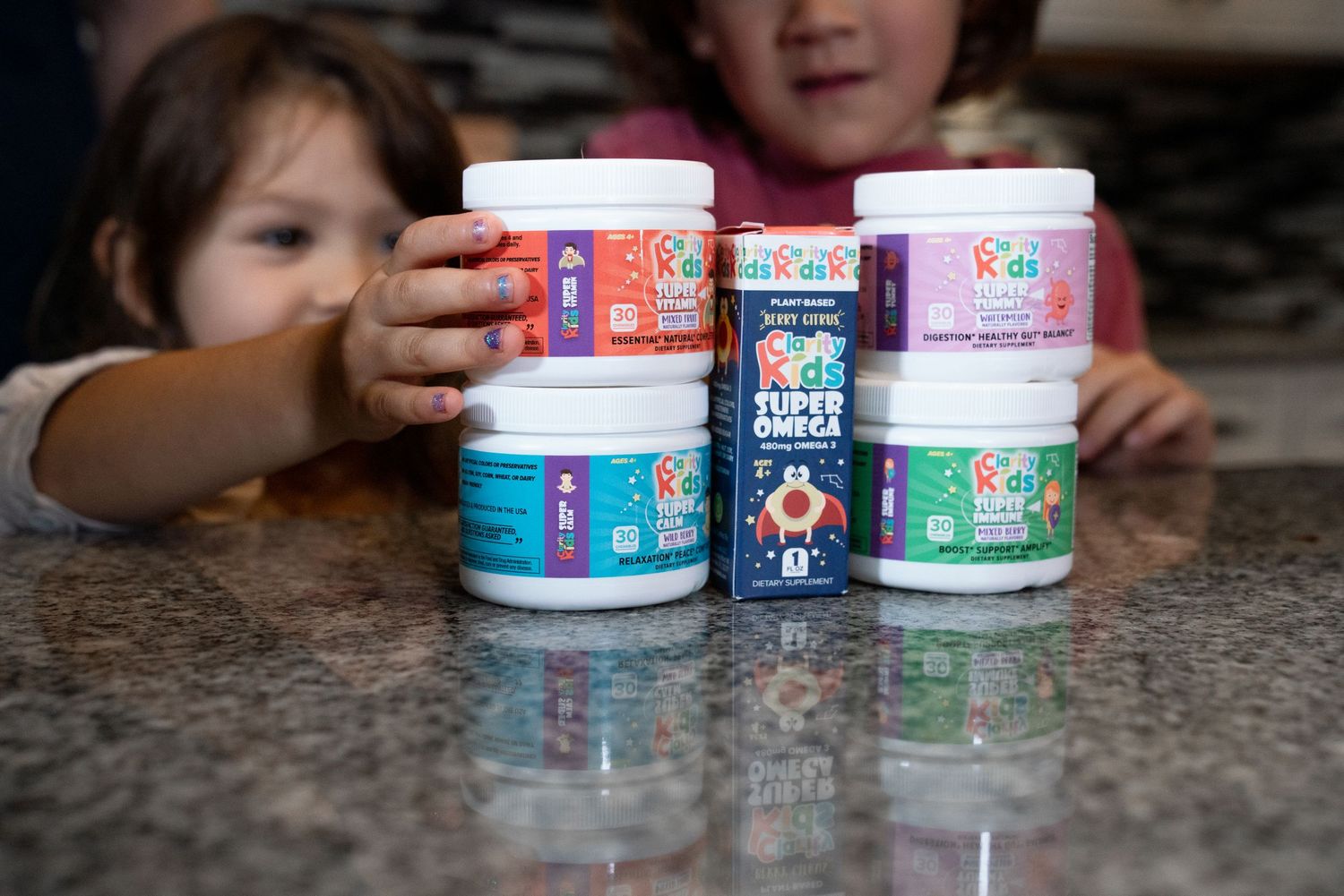Two young children reaching for multiple Kids R Kids vitamin supplement jars on a granite counter.
