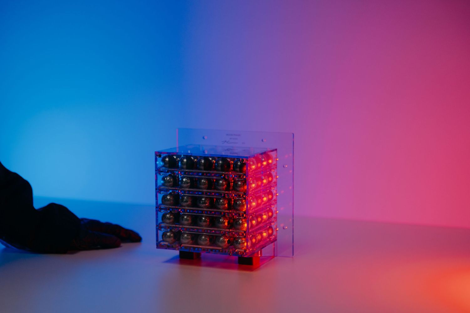 A glowing red LED cube circuit board display in a clear acrylic case under blue and pink studio lighting.