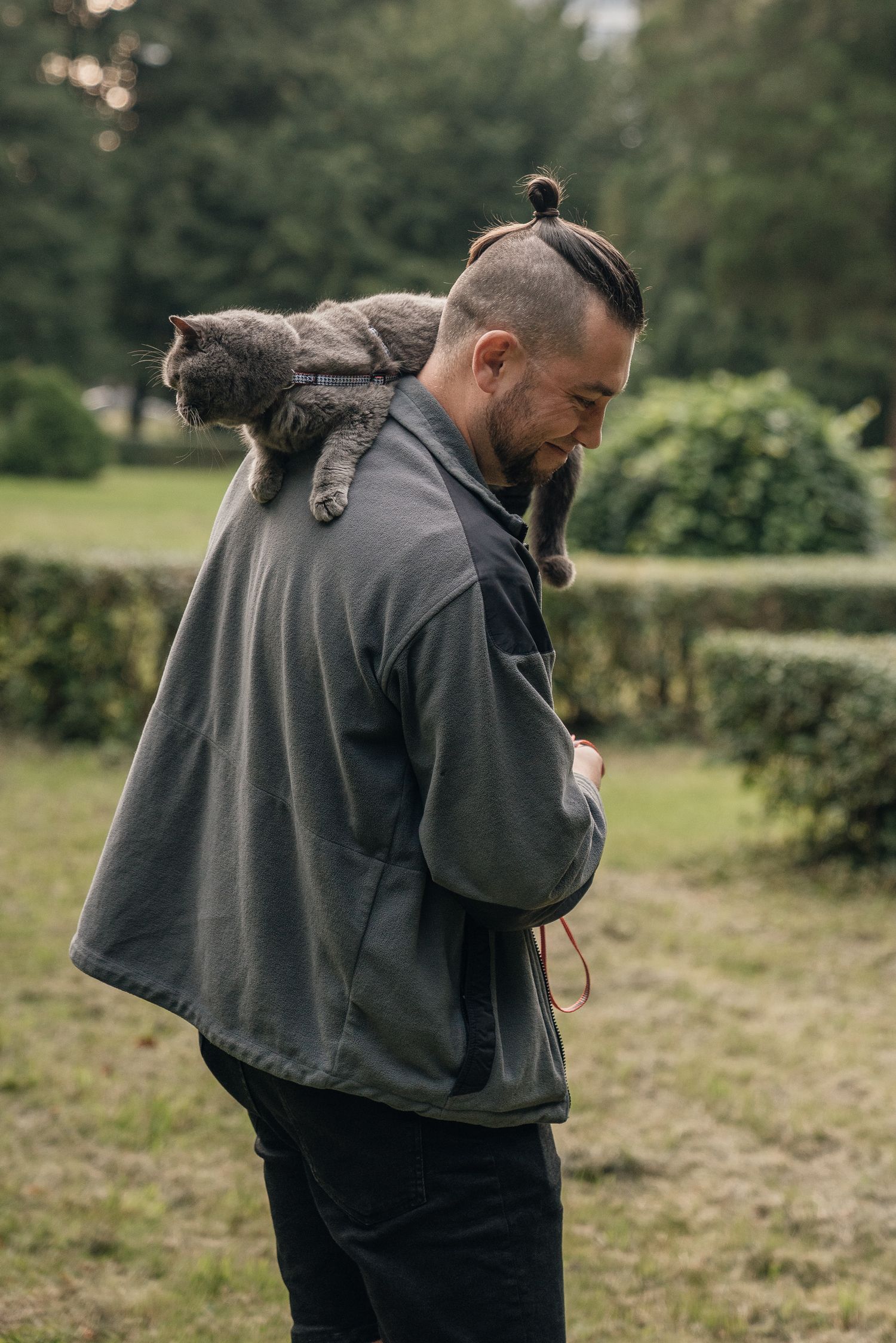 Man with top knot hairstyle and beard carries a gray tabby cat on his shoulder outdoors in a green park.