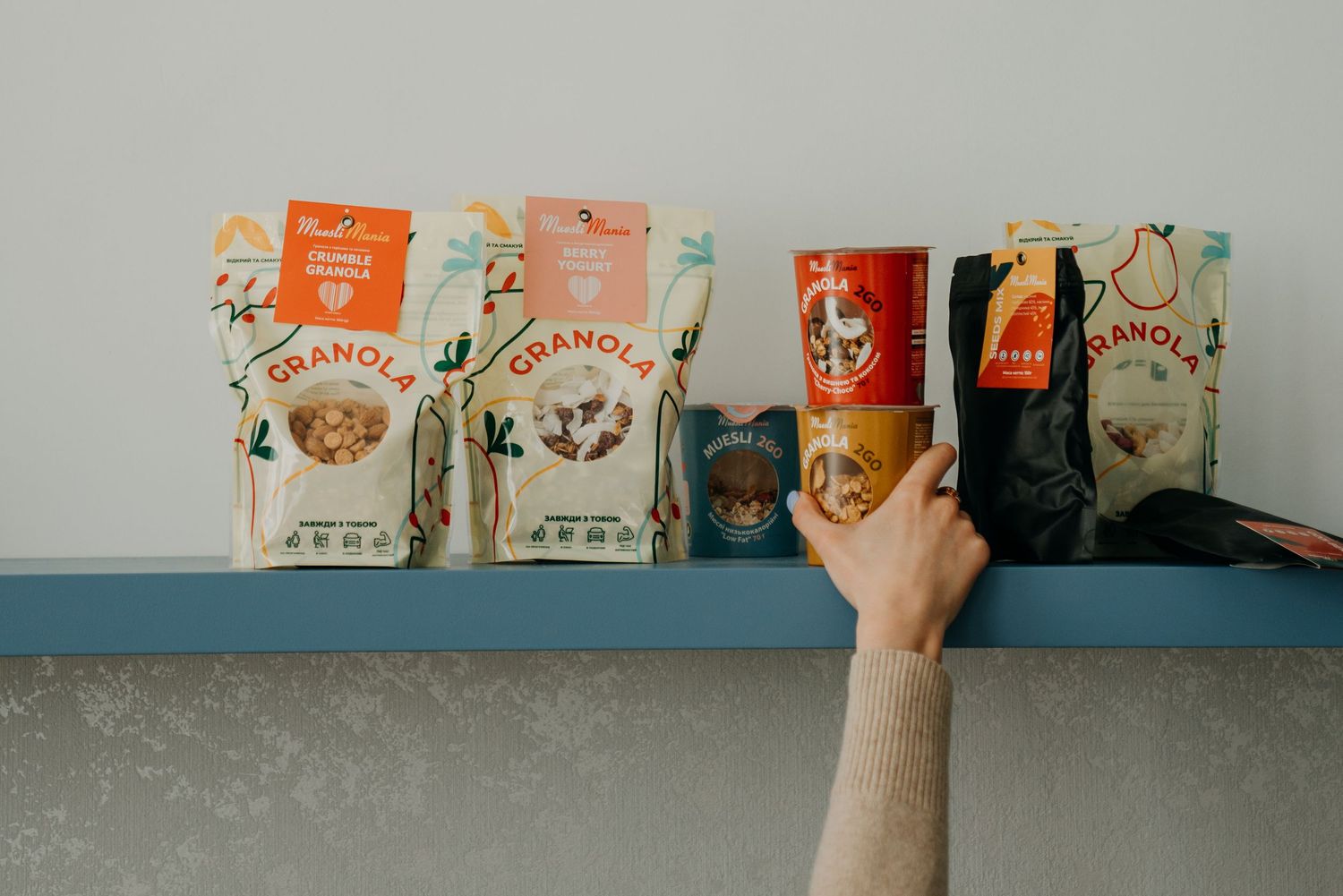 A hand reaches for colorful granola bags and cans displayed on a blue shelf against a gray wall.