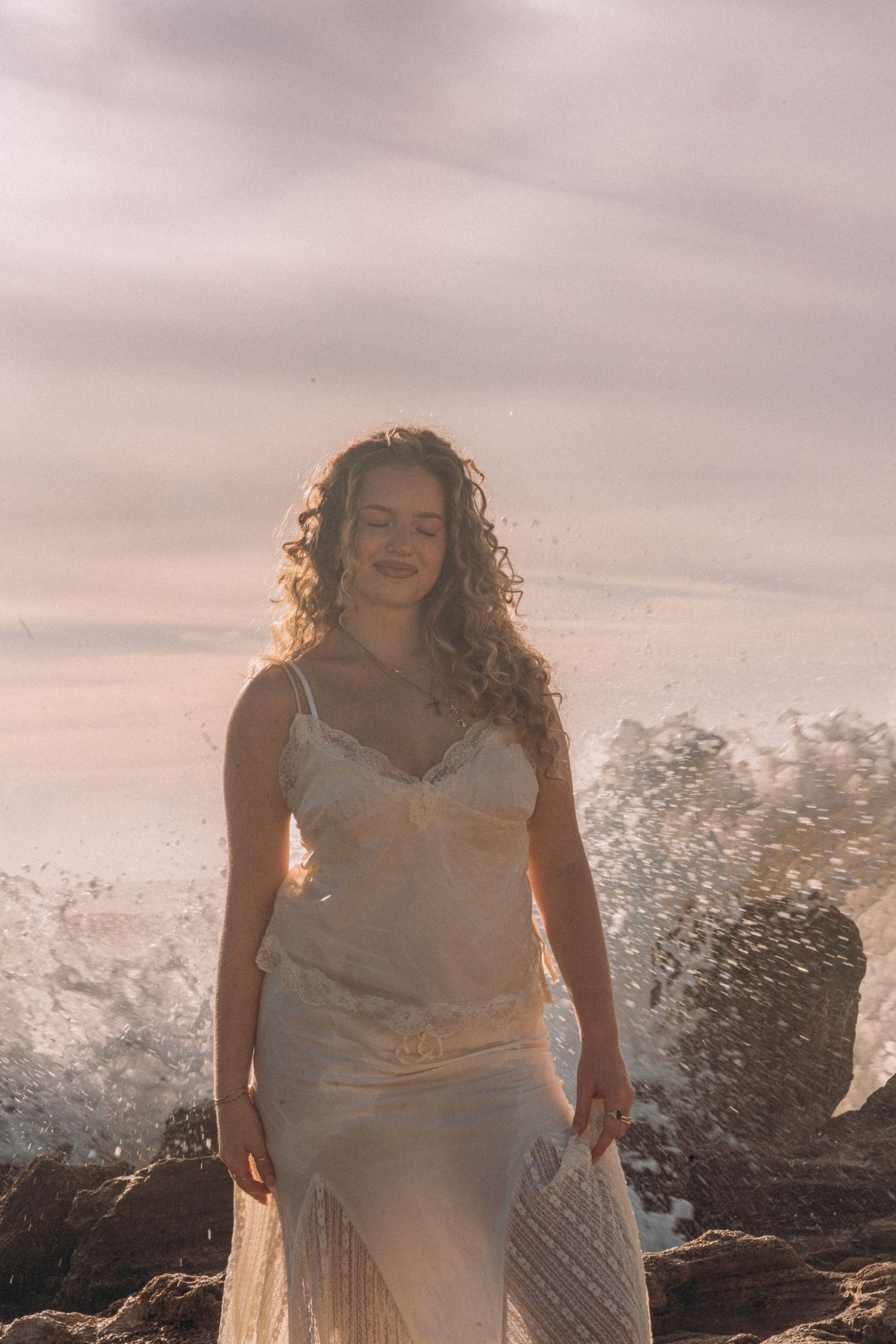Curly-haired woman in white dress smiling with ocean waves splashing behind her.