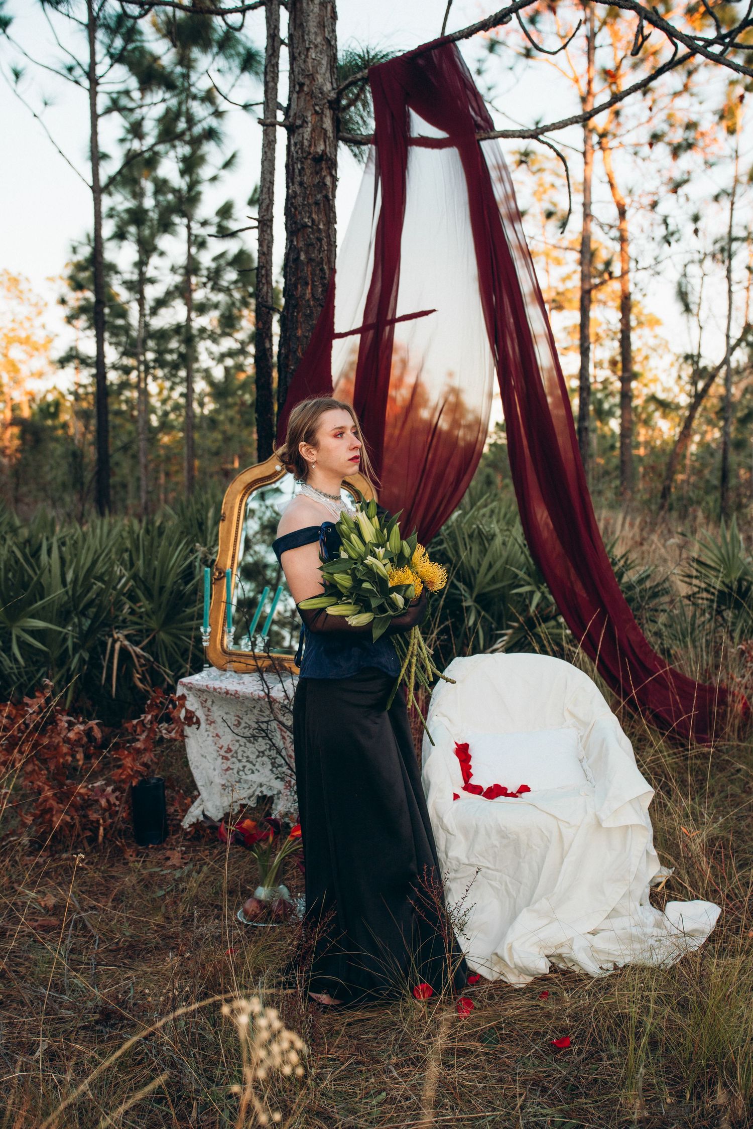 Woman in black gown holds yellow bouquet near burgundy drapes and gold mirror in forest setting.