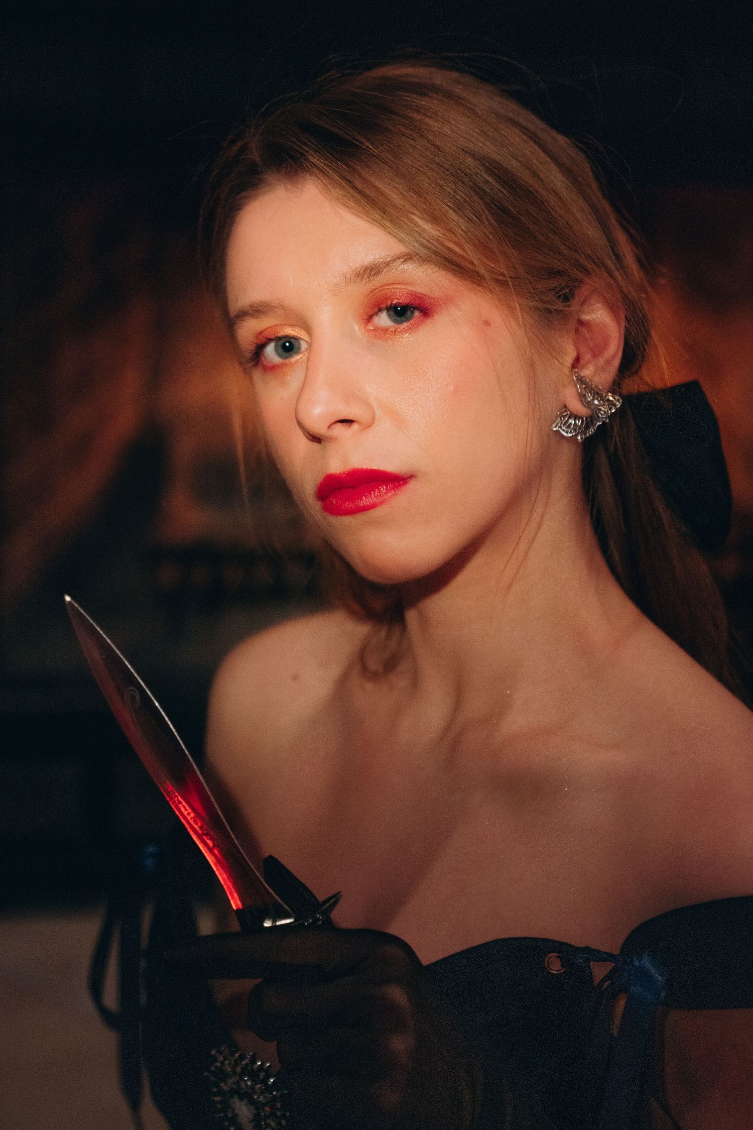 Young woman in black corset holding a bloody knife with dramatic red makeup in dark gothic setting.
