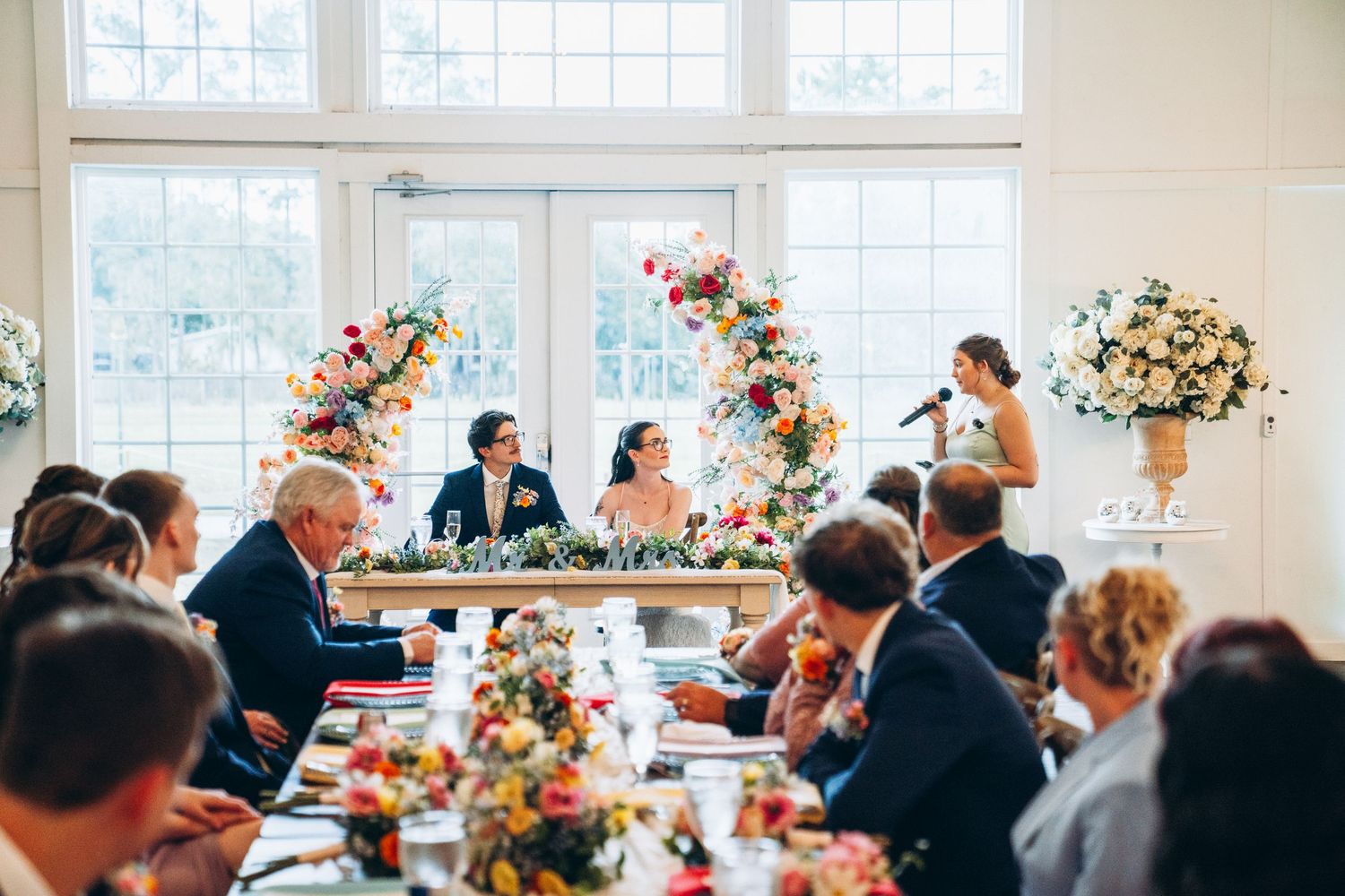 Elegant wedding reception with floral arrangements, guests seated at long tables, and a speaker addressing the bridal party.
