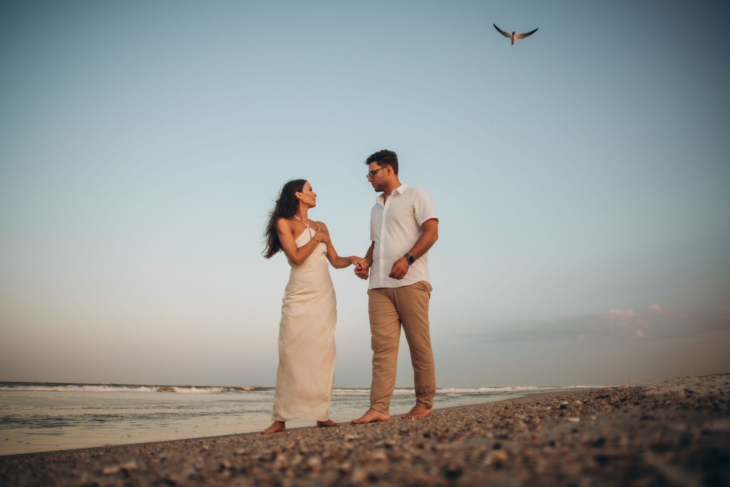 Couple holding hands on a beach at sunset with a bird flying in the clear sky behind them.