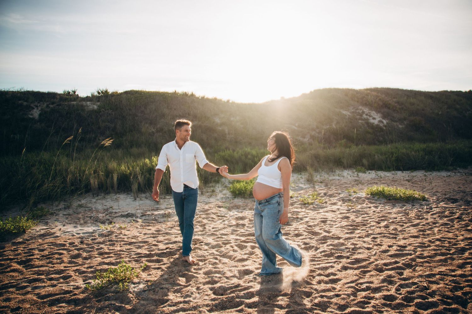 Couple holding hands and walking on sandy beach dunes at golden sunset, man in white shirt and woman in casual attire.