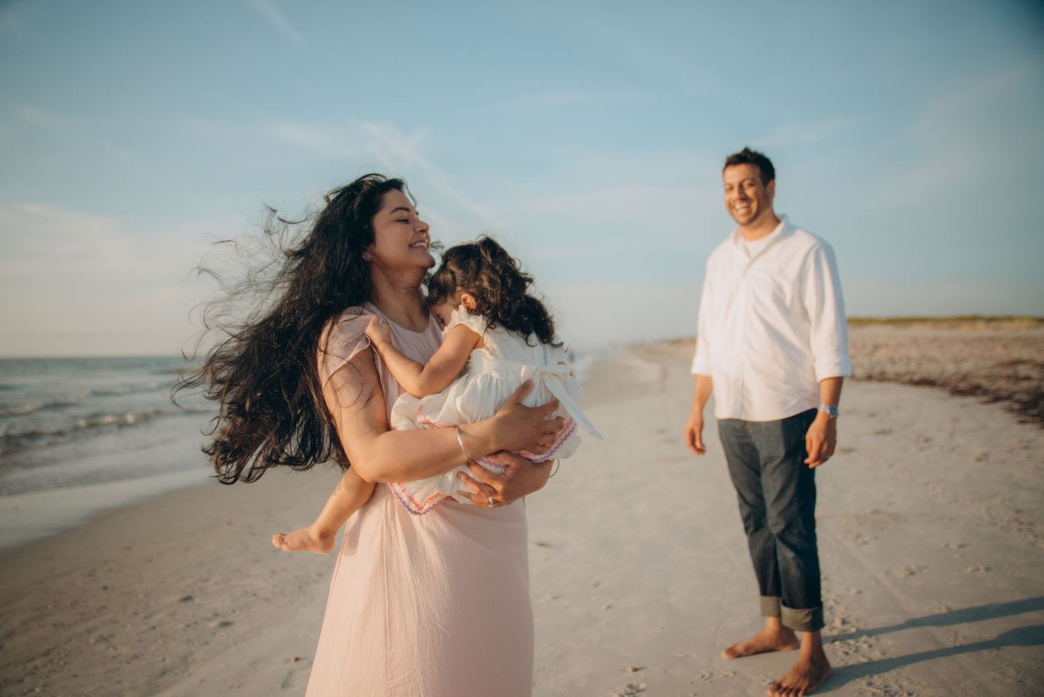 Family beach photo with mother holding baby girl, father smiling in background, ocean shore, golden hour light.