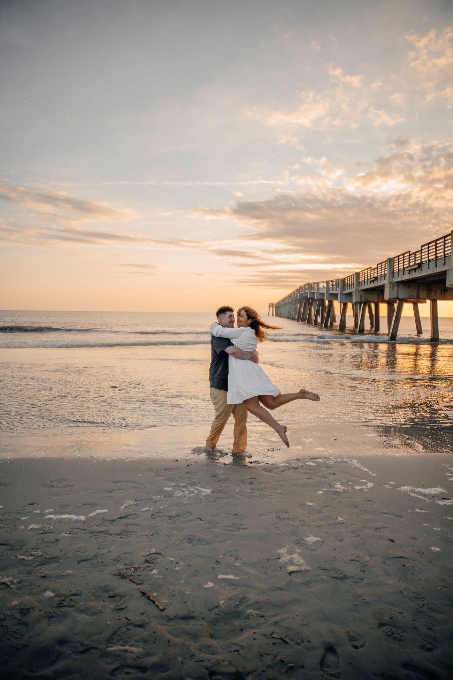 A playful couple dances on the beach near a wooden pier during sunset.