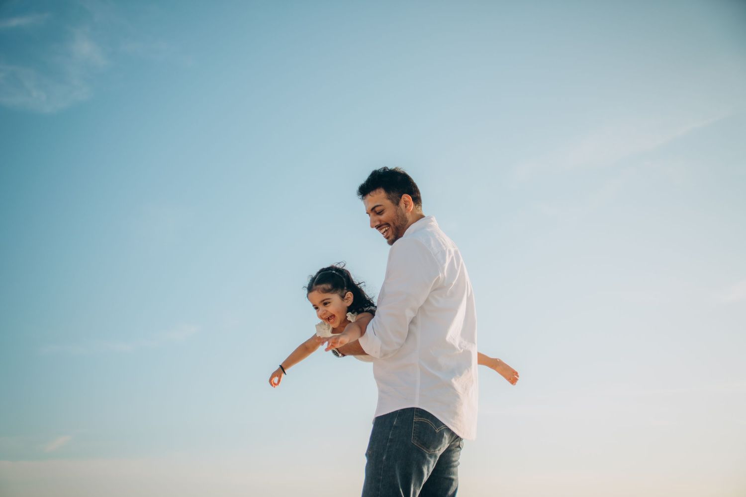 A smiling father joyfully lifts his happy child outdoors against a bright blue sky.