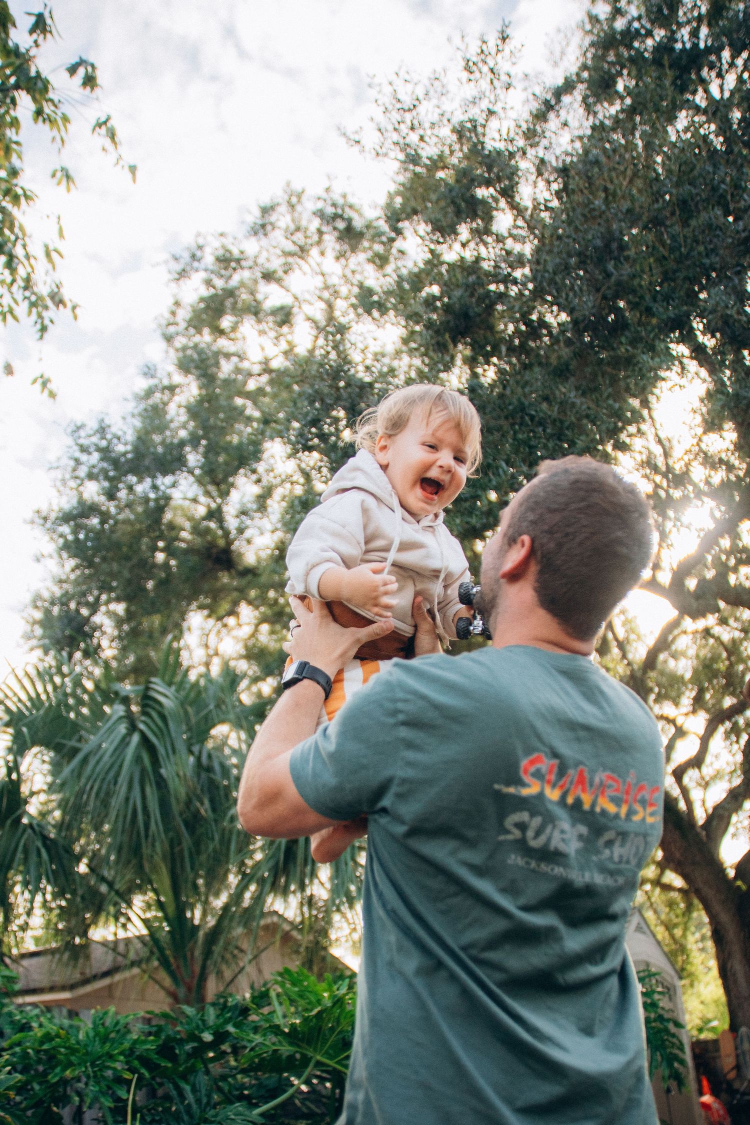 A person in green lifts a small child playfully in the air during a sunny afternoon.