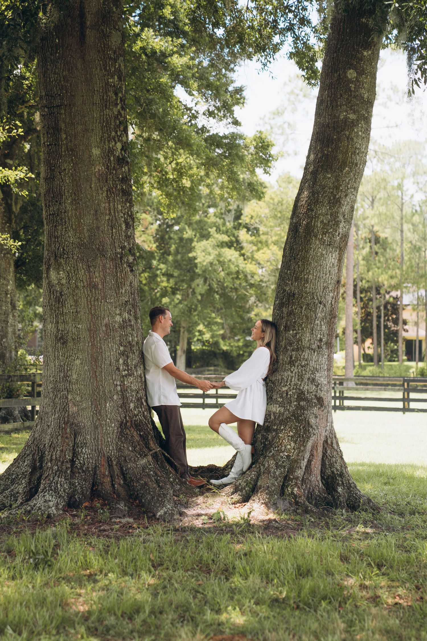A couple poses playfully between two massive oak trees on a sunny afternoon.