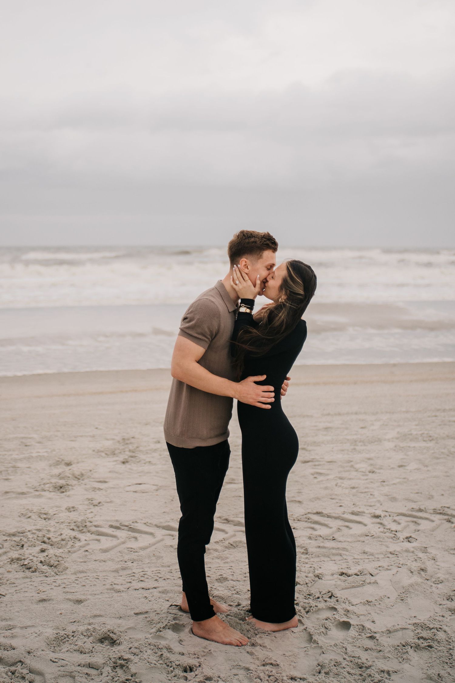 A couple shares a romantic embrace while standing barefoot on a sandy beach.