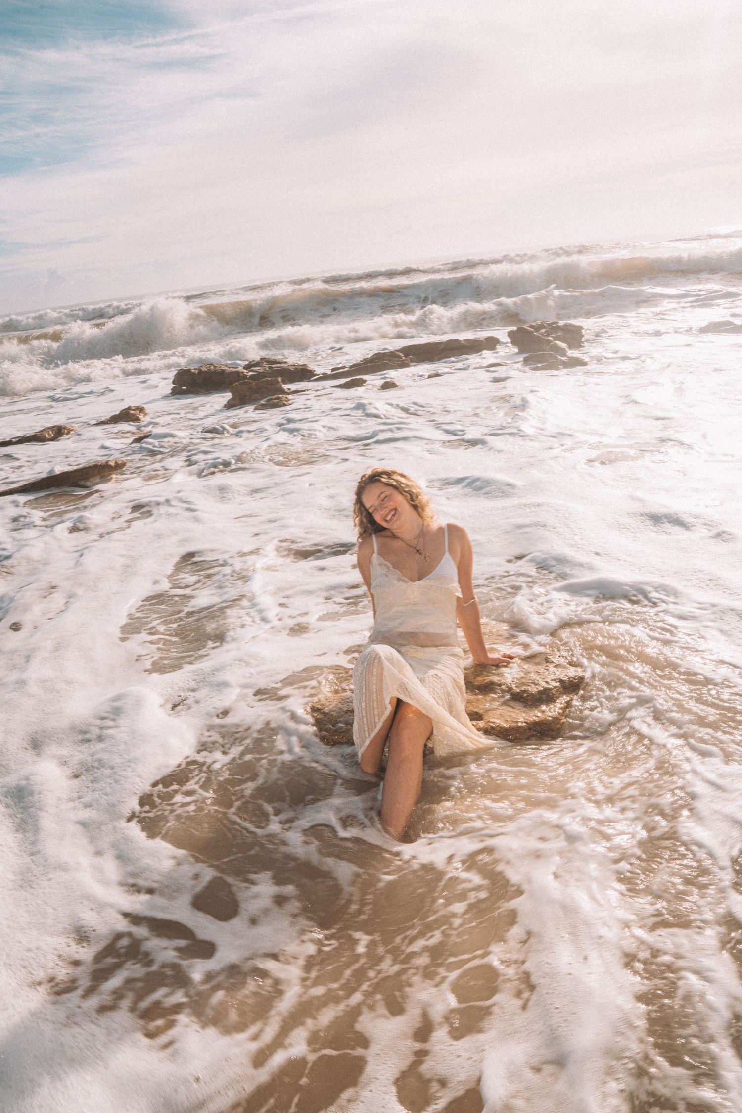 Woman in white dress sitting on rocks as ocean waves wash over her on a sunny beach.