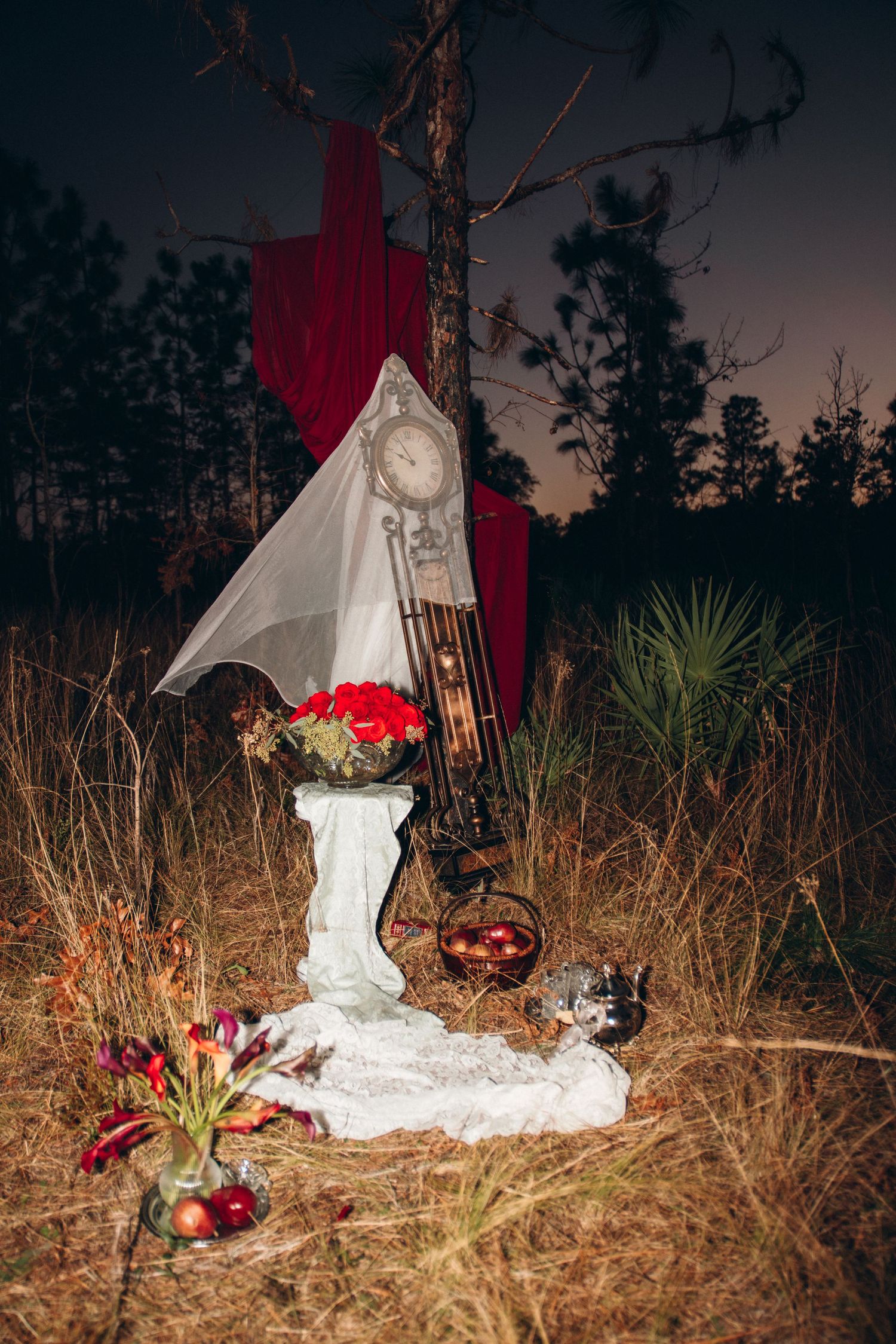 Mystical outdoor night scene with vintage clock, white pedestal, red roses, and dramatic crimson draping in forest.