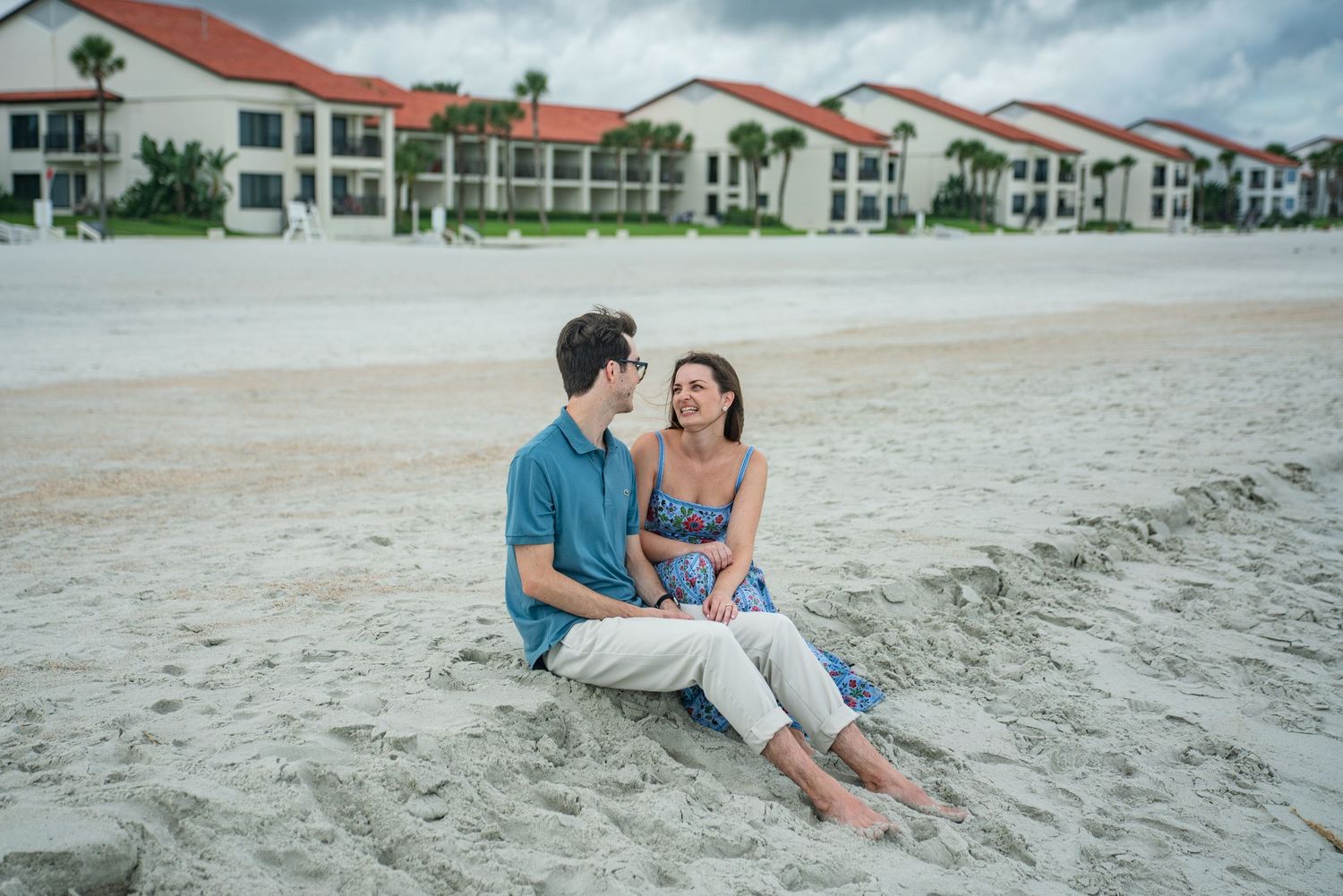 A couple sits together on a sandy beach with white coastal condominiums in the background.