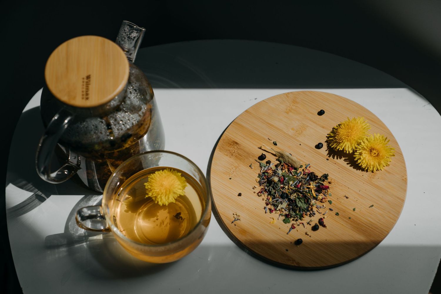 Glass teapot and cup with dandelion flower tea on white table with wooden board and loose herbs.