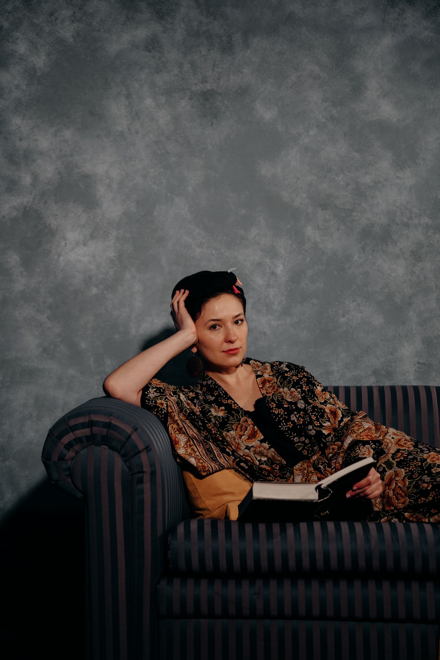 Woman in floral dress reclines on striped sofa holding book, posing thoughtfully against gray textured studio backdrop.