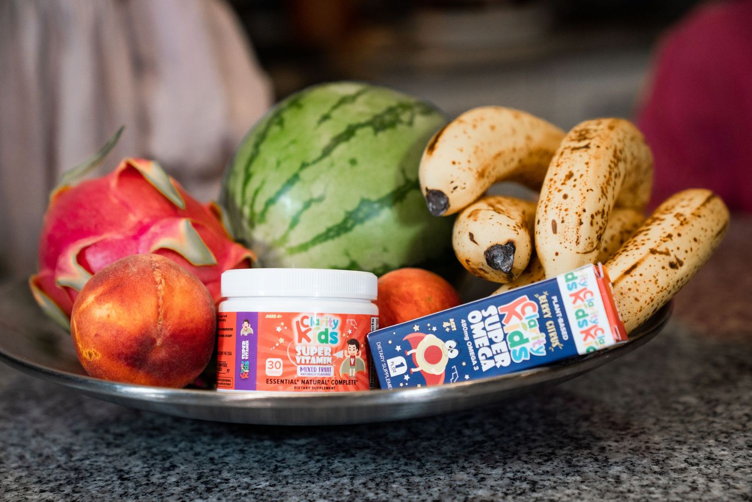 A metal bowl filled with fresh fruits including watermelon, bananas, peach, dragon fruit, and vitamin supplement bottles.