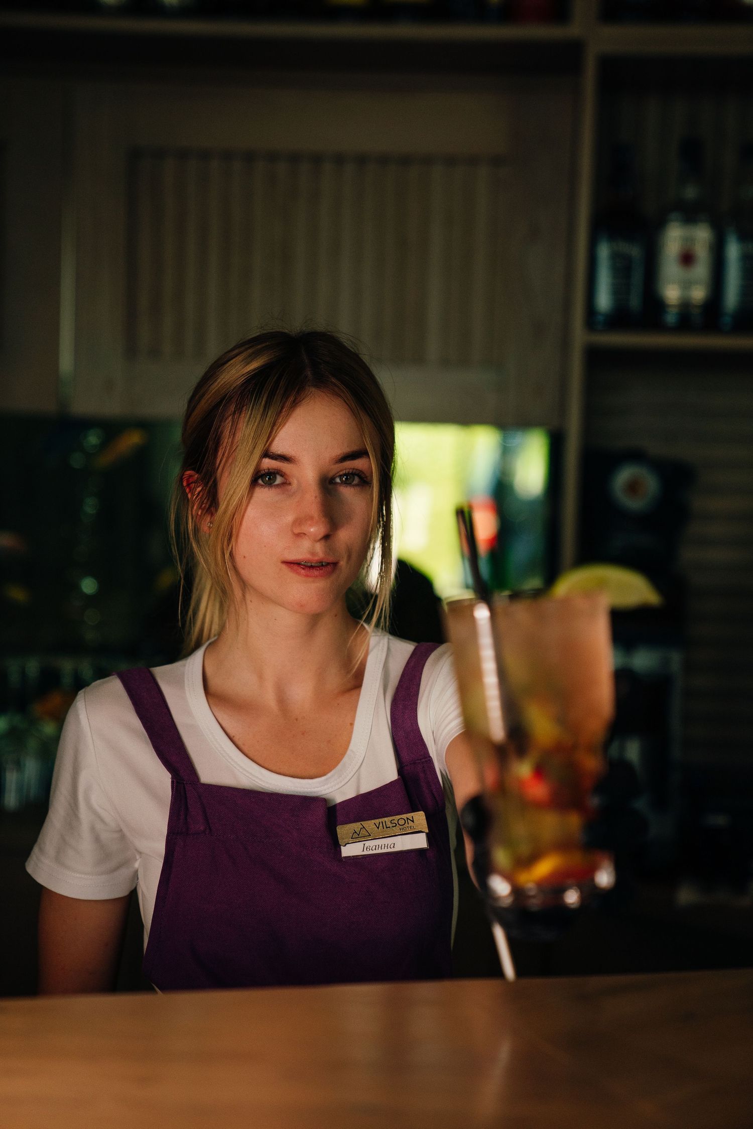 Female bartender in purple apron presenting a tall tropical cocktail at a bar counter.