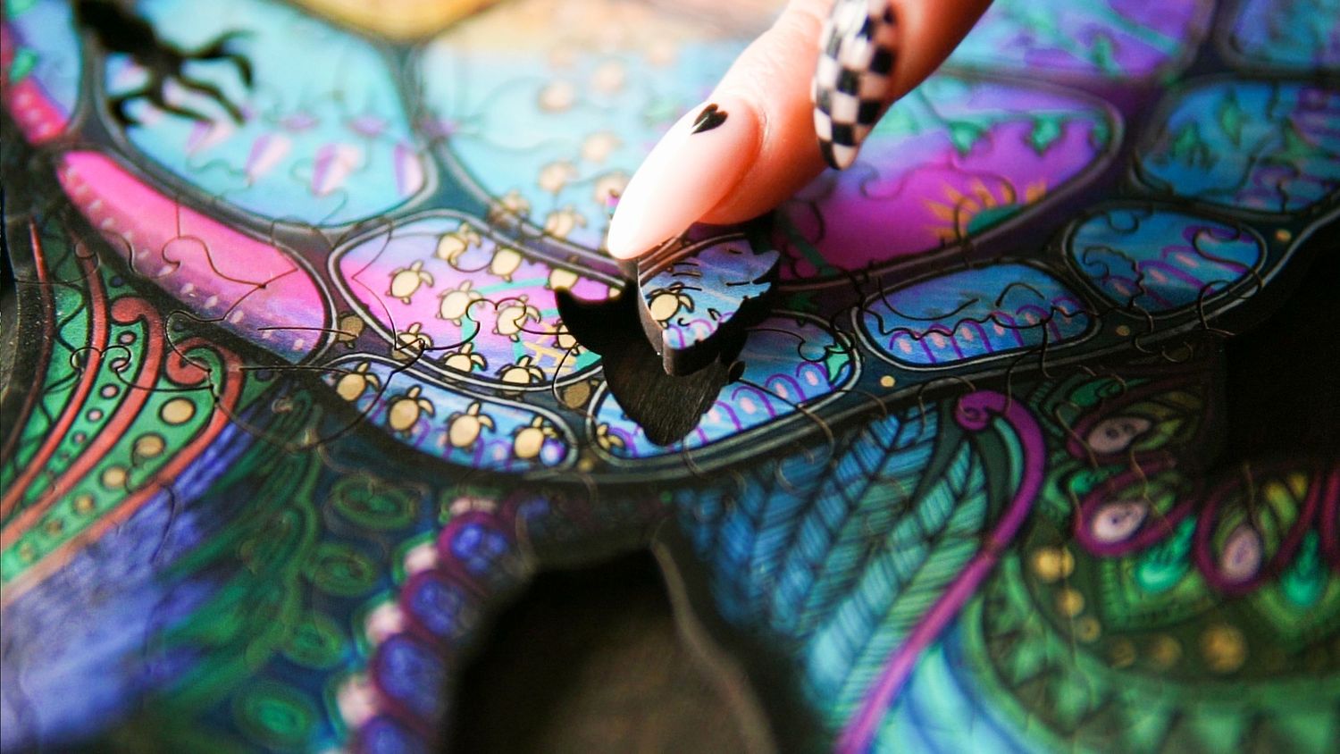 A hand with a decorative nail holds a pen over a vibrant, colorful mandala artwork with intricate patterns.