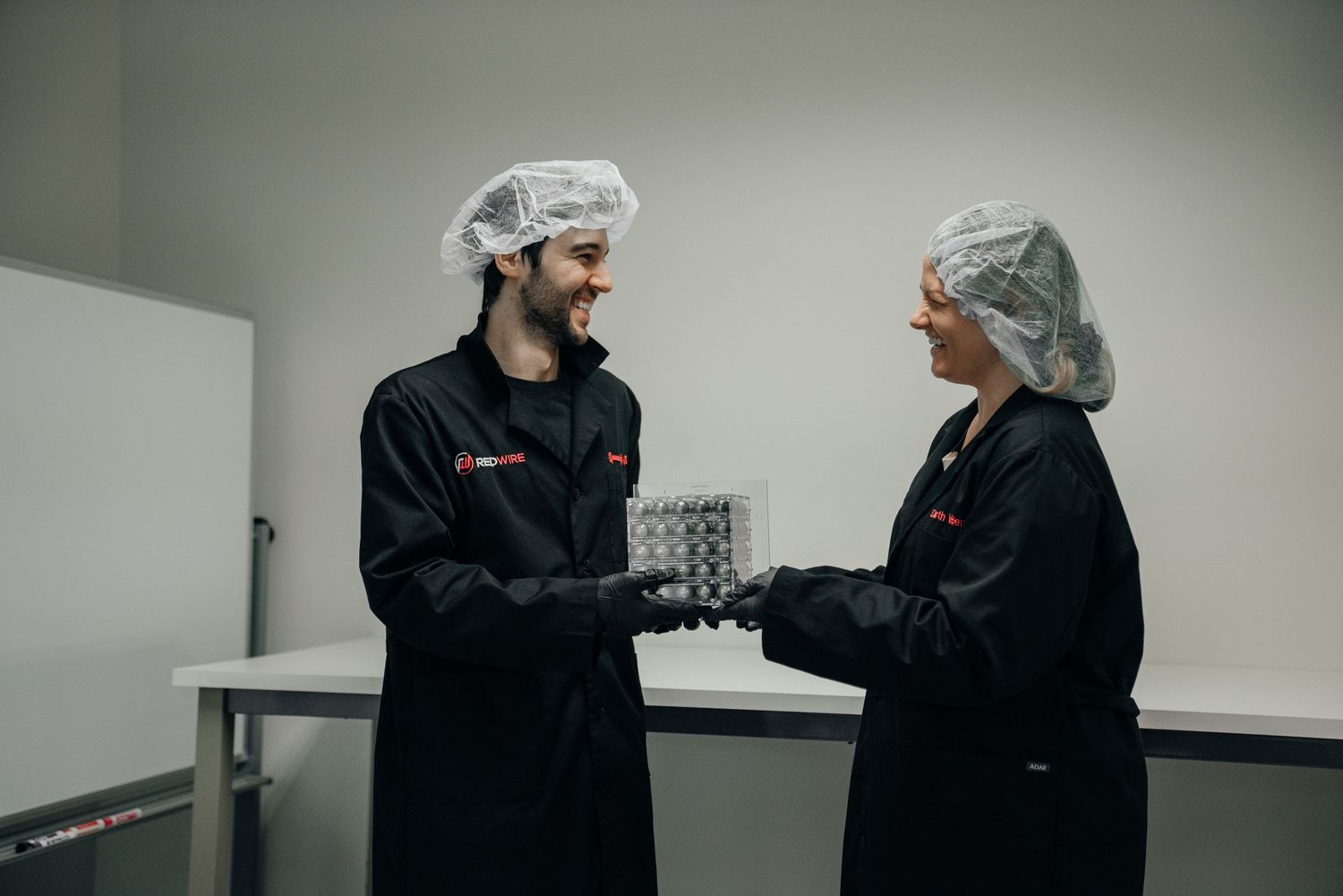 Two lab workers in black uniforms and hair nets exchange a sample tray in a clean laboratory environment.