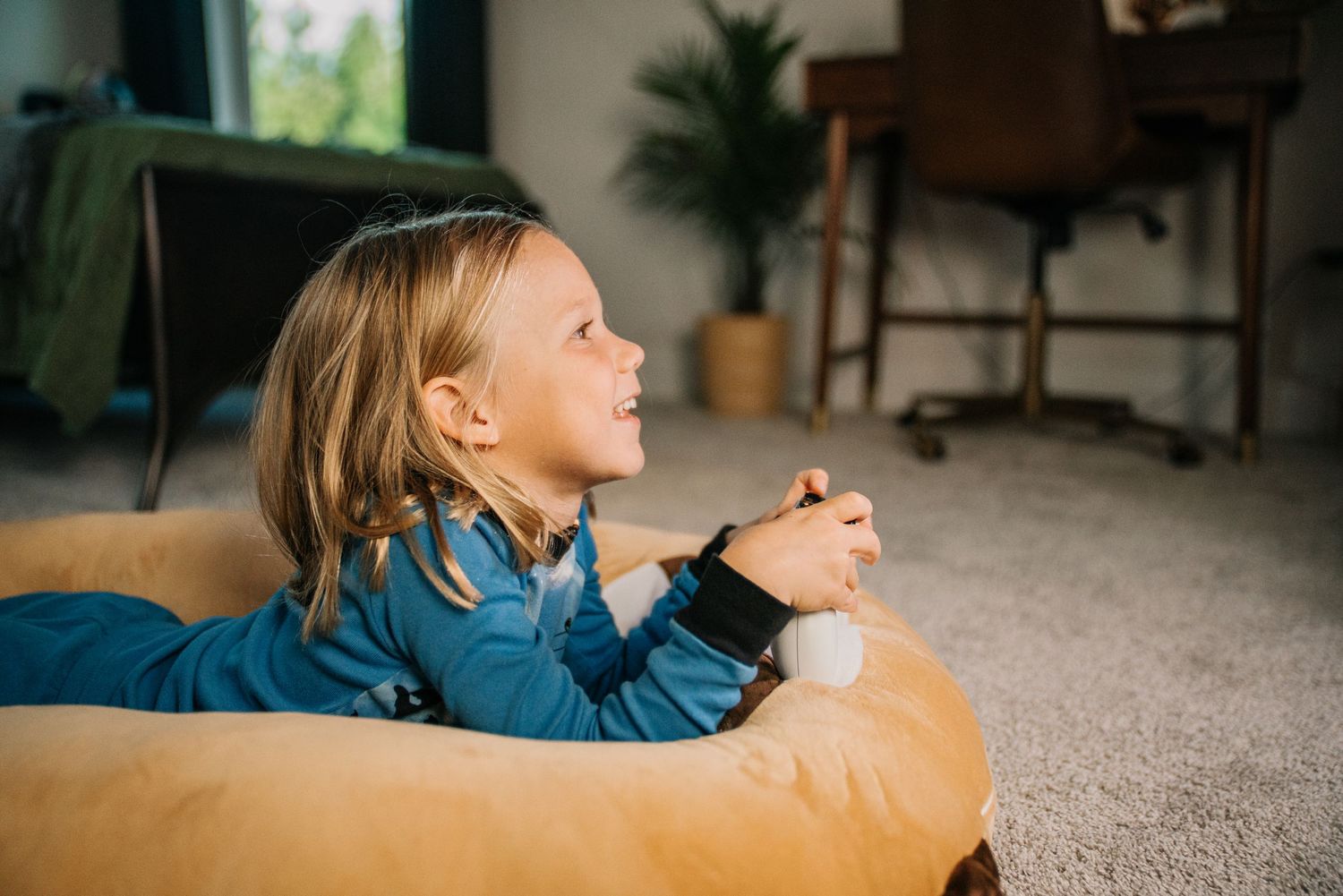 Cheerful child relaxing on a tan bean bag pillow on carpet, looking up with a smile in a cozy room.