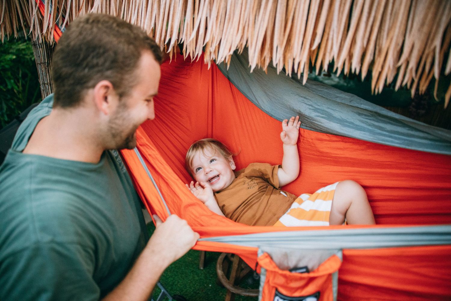 Family enjoying quality time together on a bright orange hammock under a thatched canopy.