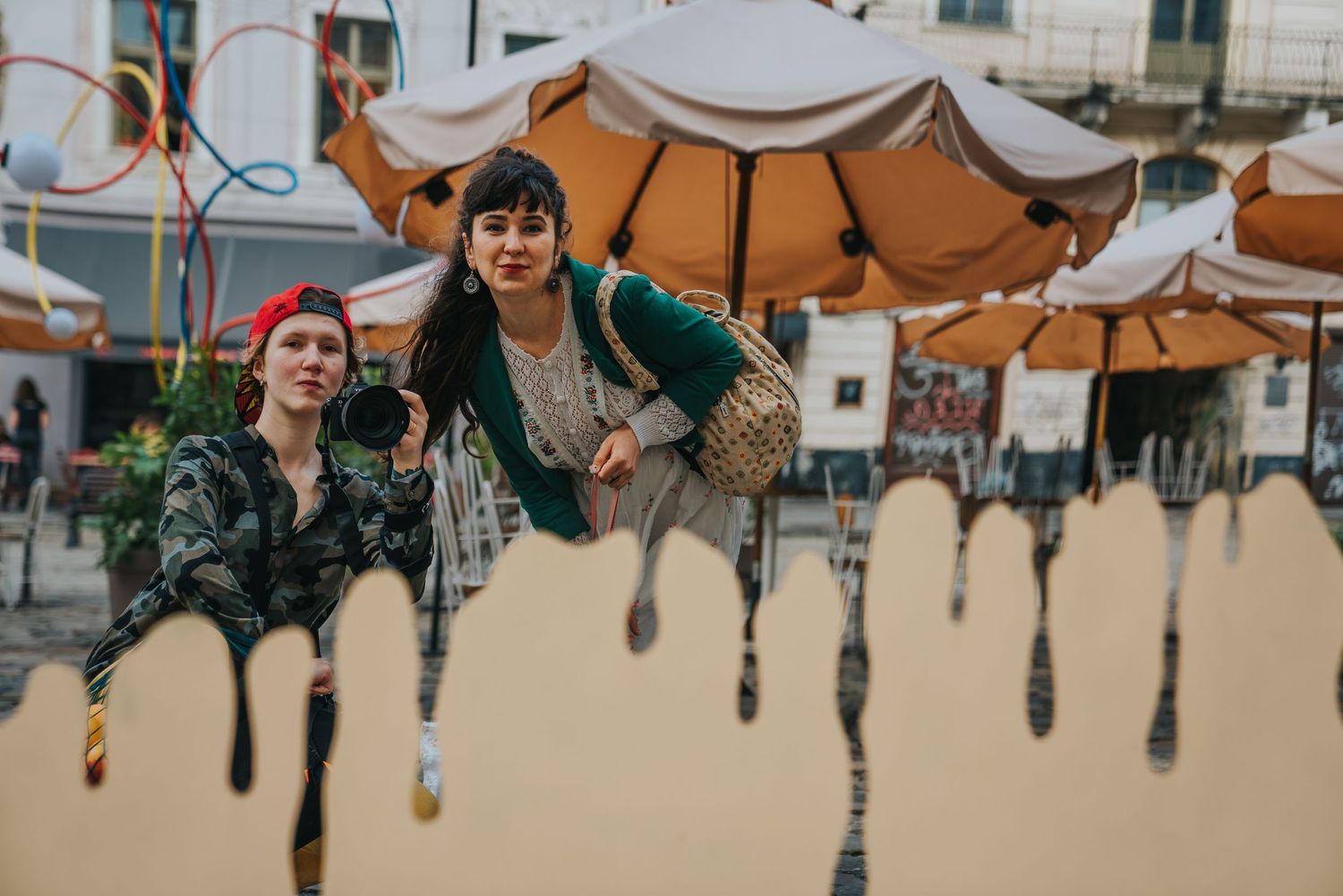 Two women pose playfully behind a dripping paint art installation outdoors near cafe umbrellas.