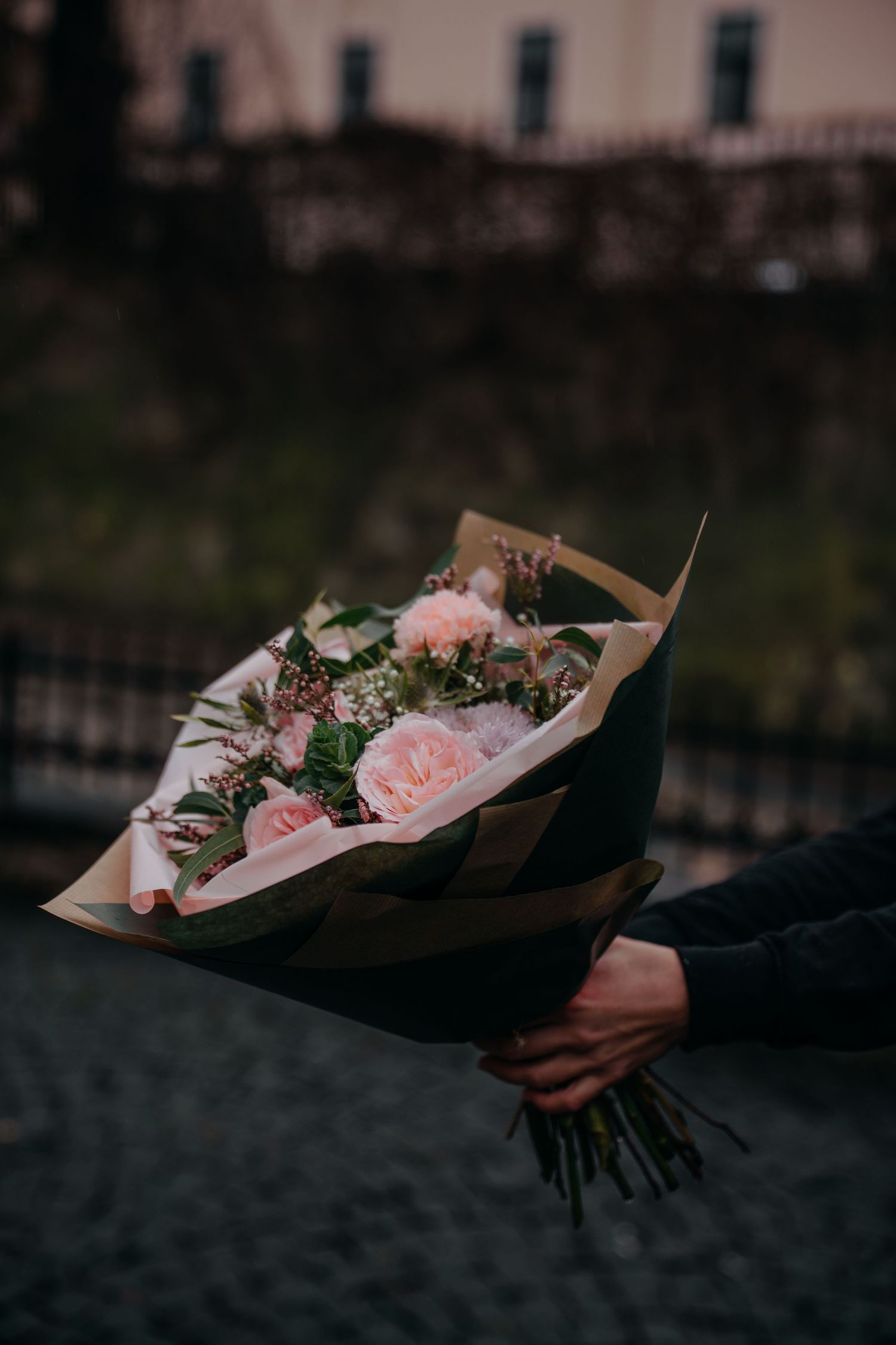 Hand holding an elegant bouquet of pink roses wrapped in kraft paper outdoors.