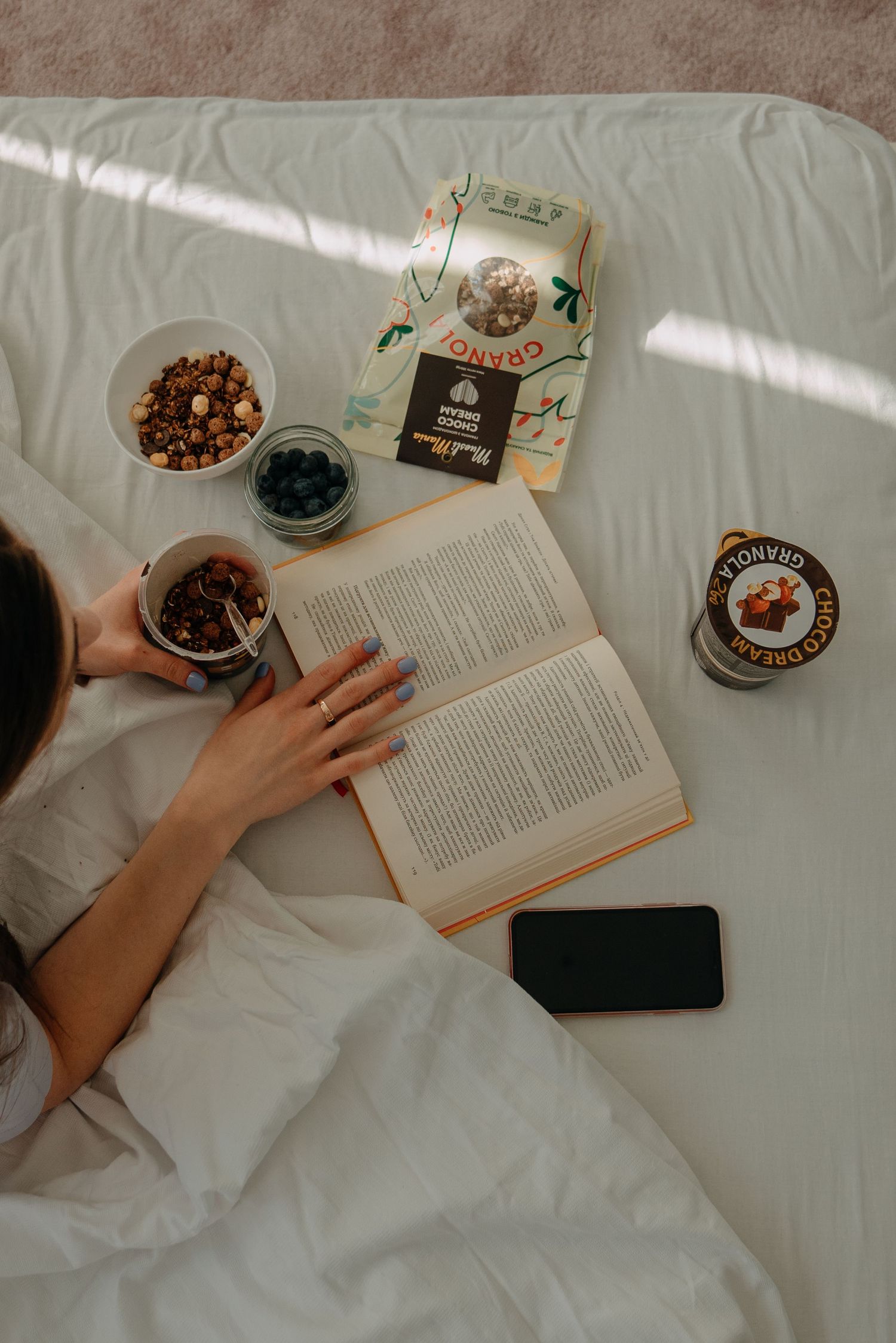 Woman reading a book in bed with granola snacks, blueberries, and a smartphone on white bedding in morning light.