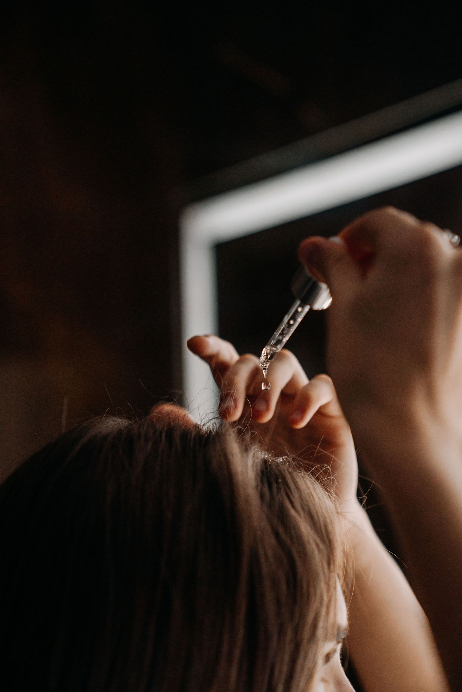 A person applying hair serum with a dropper to their scalp, with a lit mirror in the background.