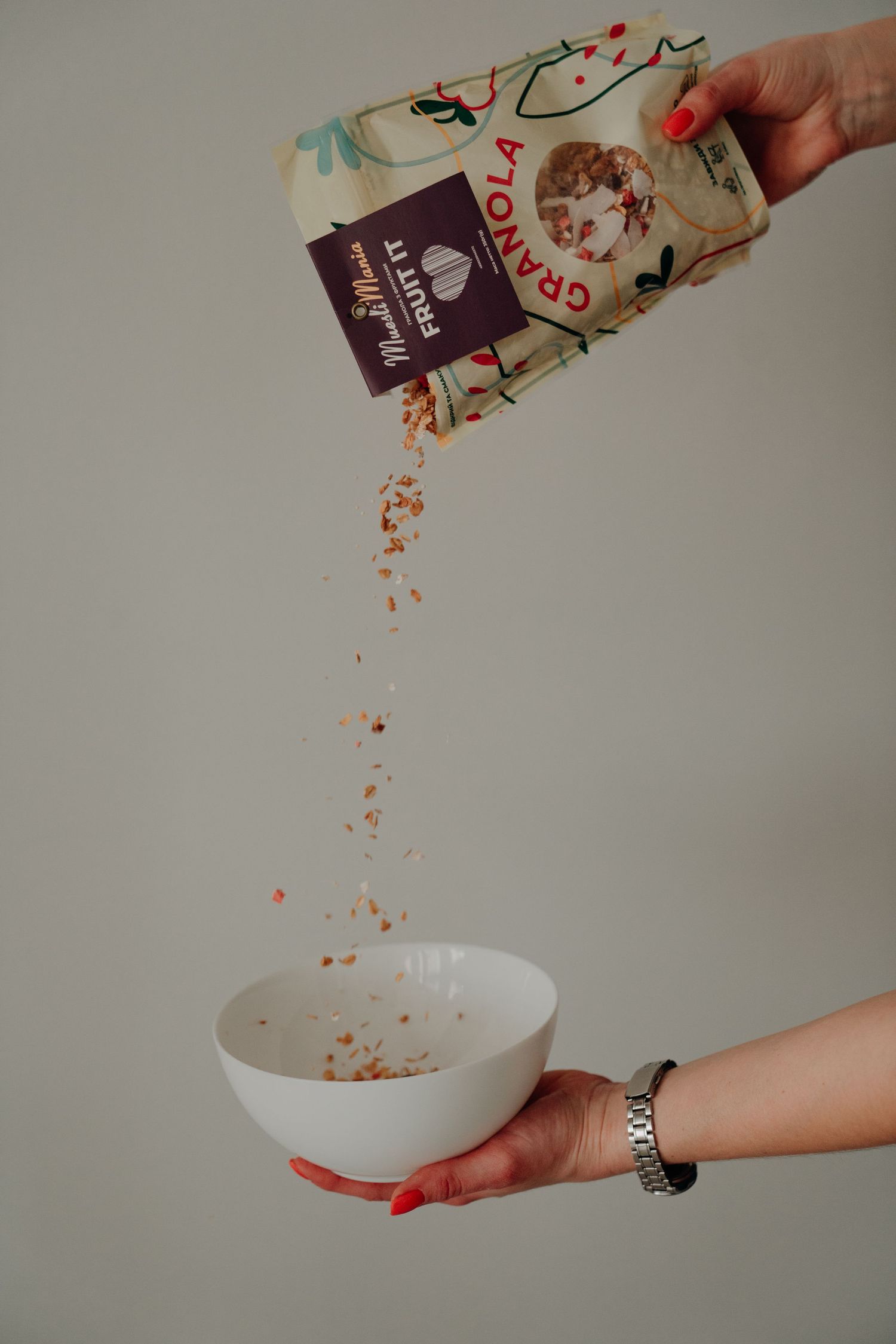 Hand with red nails pouring granola from a MuesliMania bag into a white bowl.