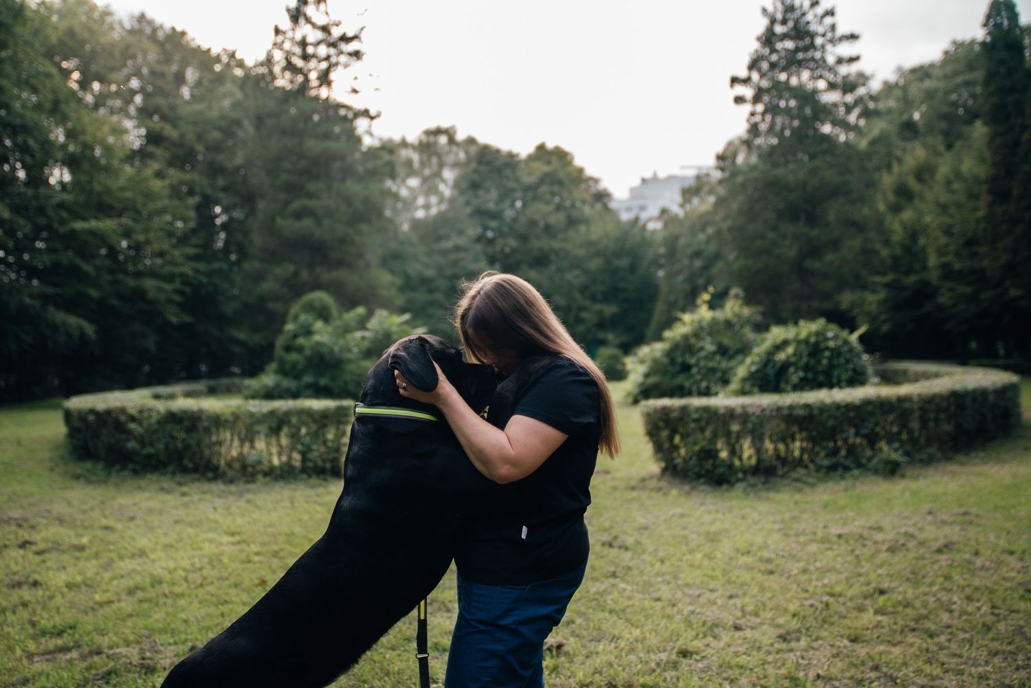 Woman in black outfit hugging a large black dog in a lush green park with trimmed hedges.