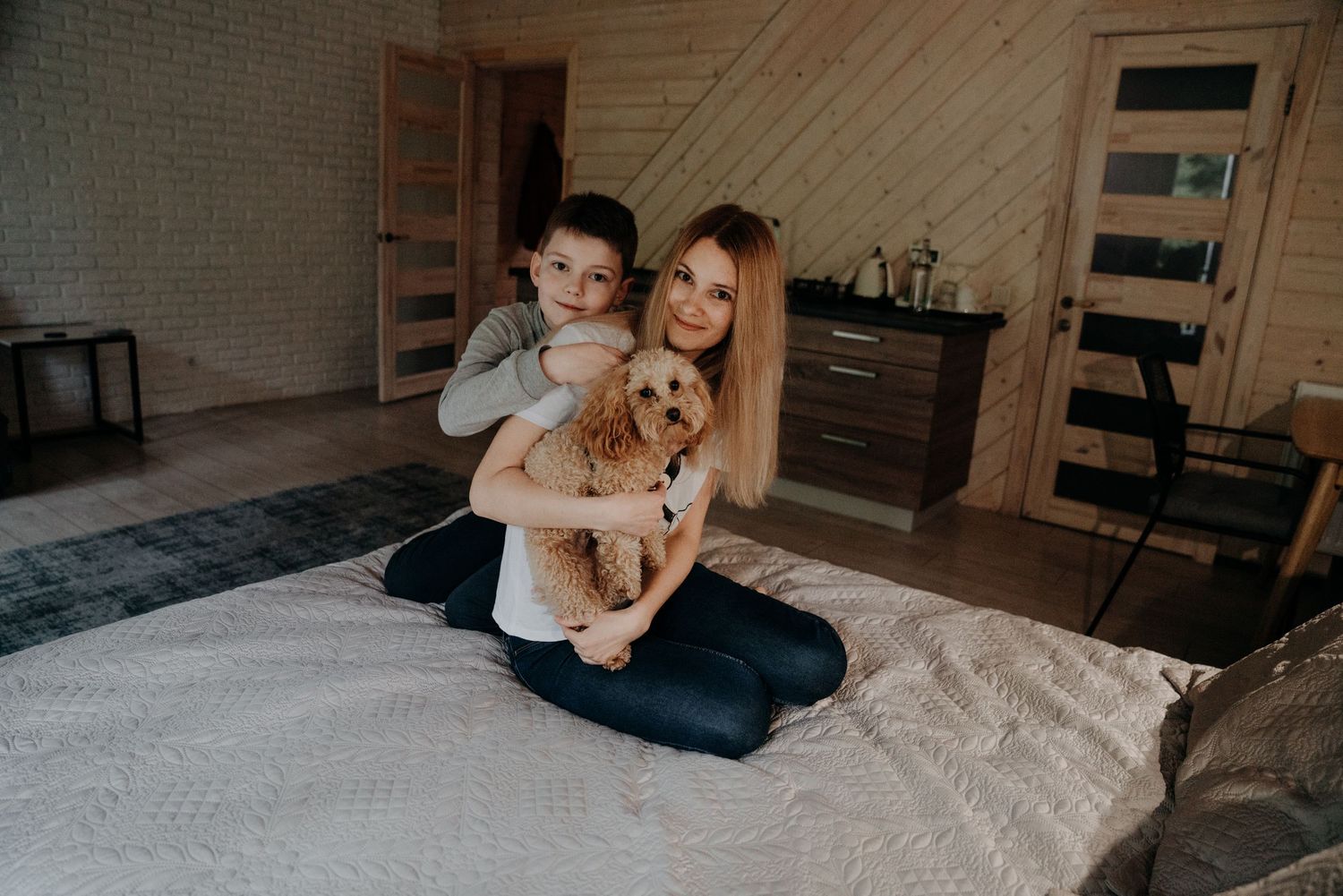 Woman and boy sitting on bed hugging a fluffy brown dog in a cozy wooden bedroom.
