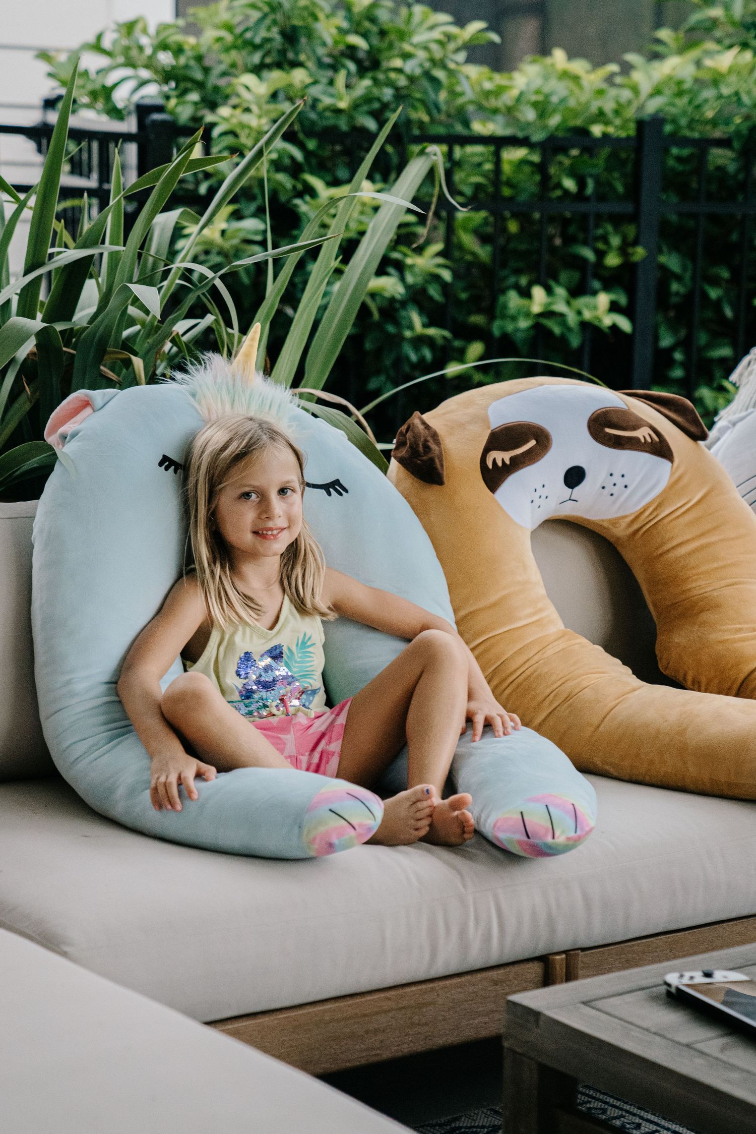 Young girl smiling on outdoor sofa with large unicorn and sloth animal plush pillows surrounded by greenery.