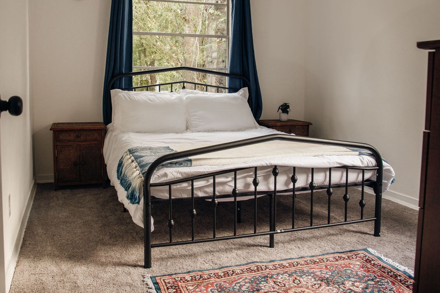 Cozy bedroom with black iron bed frame, white bedding, teal curtains, and a vintage rug on carpet.